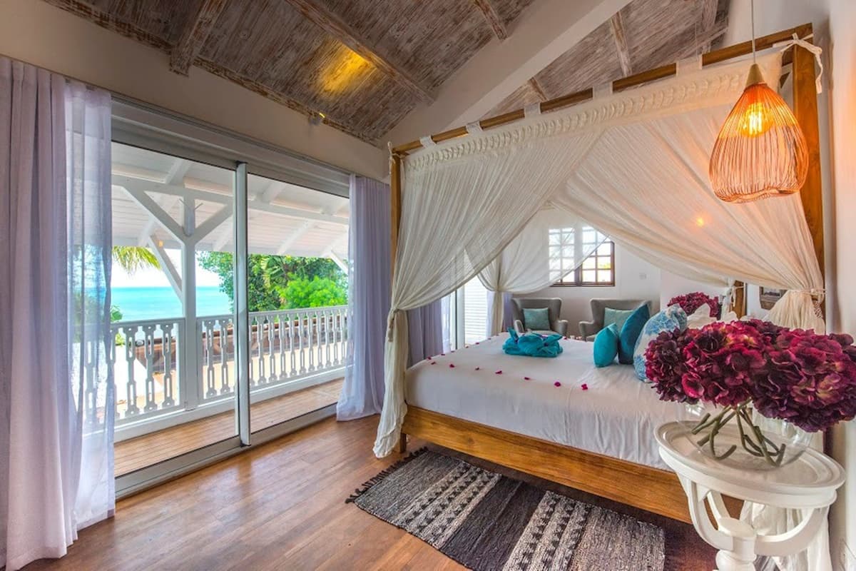 Een slaapkamer met een houten bed, een hemelbed, uitzicht op de oceaan en een balkon met een witte leuning.