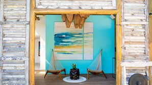 Een kamer met een houten vloer, twee rieten stoelen, een kleine tafel met een decoratief object, en een groot abstract schilderij aan de muur.