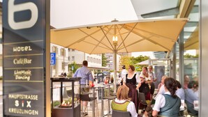 Un espacio al aire libre con mesas y sombrillas, un letrero para un hotel y café/bar, y gente comiendo y conversando.
