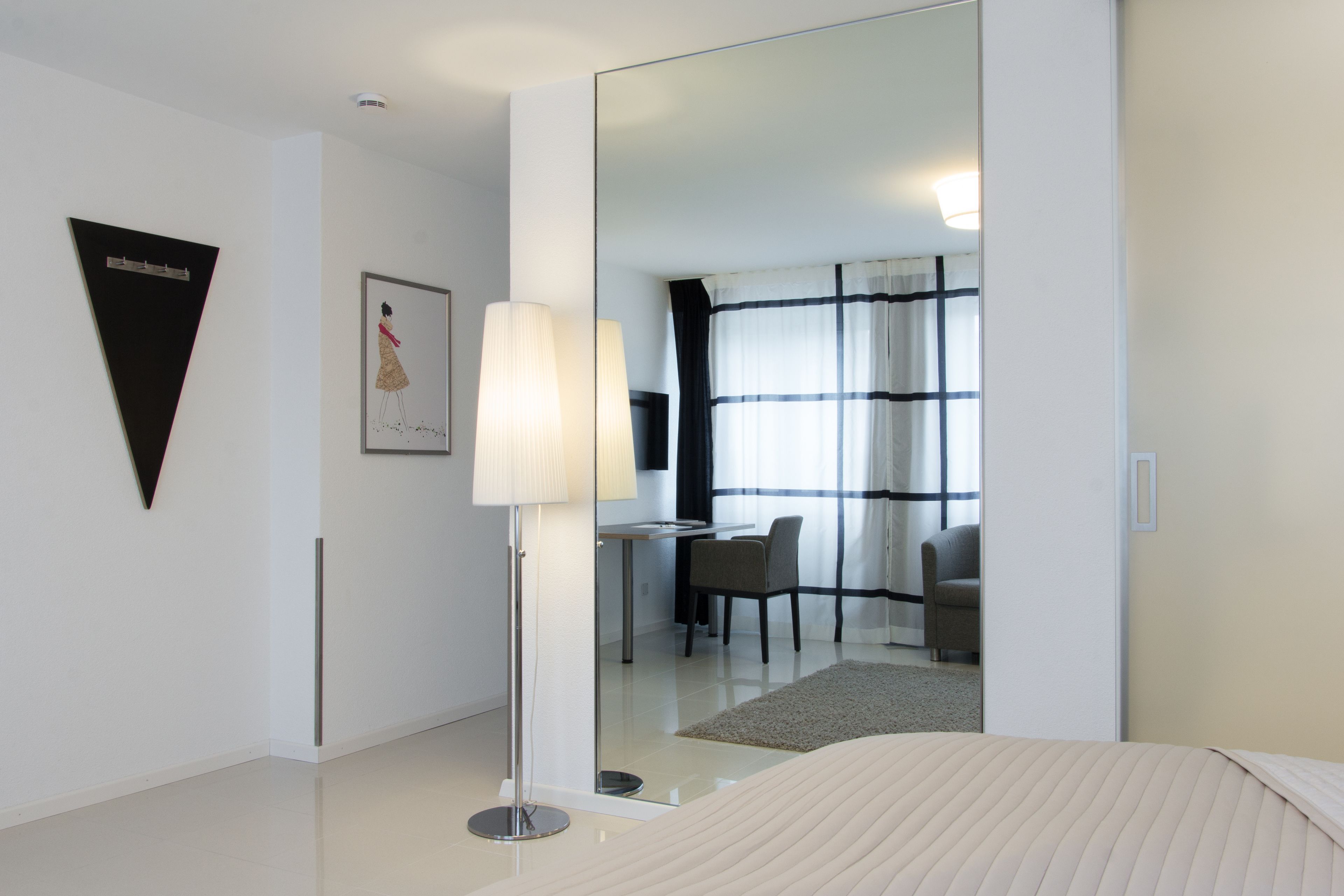A modern hotel room with a large bed, a desk with a chair, a mirror, and a floor lamp.