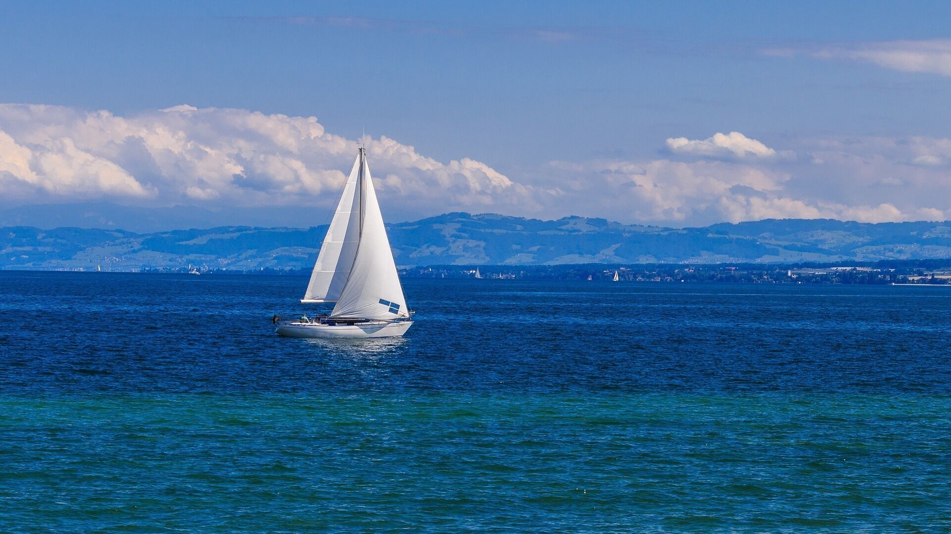 Un velero en un mar tranquilo con una costa lejana y montañas al fondo.