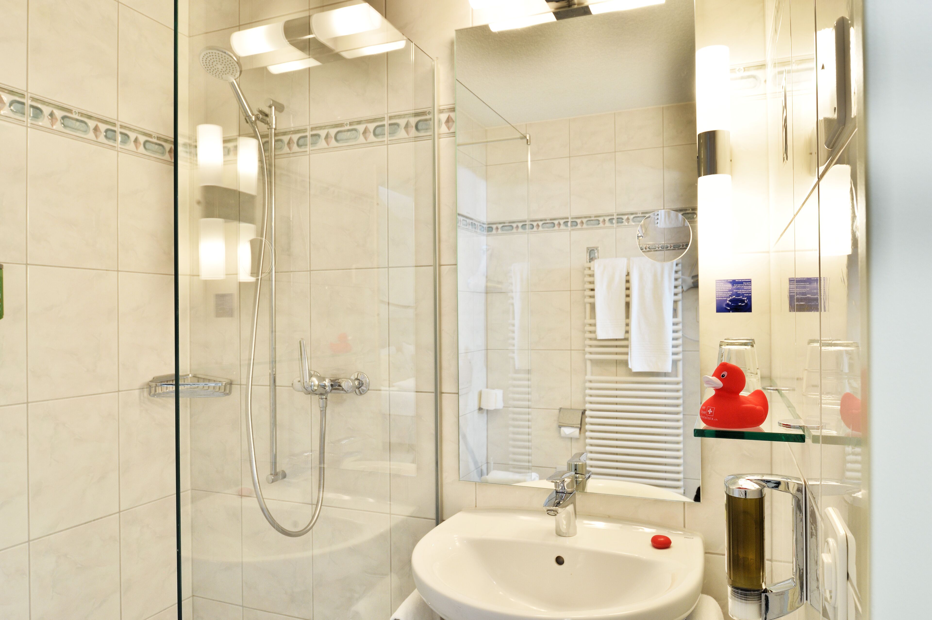 Un baño moderno con ducha envuelta en vidrio, un lavabo blanco y un estante de vidrio con un patito de goma rojo.