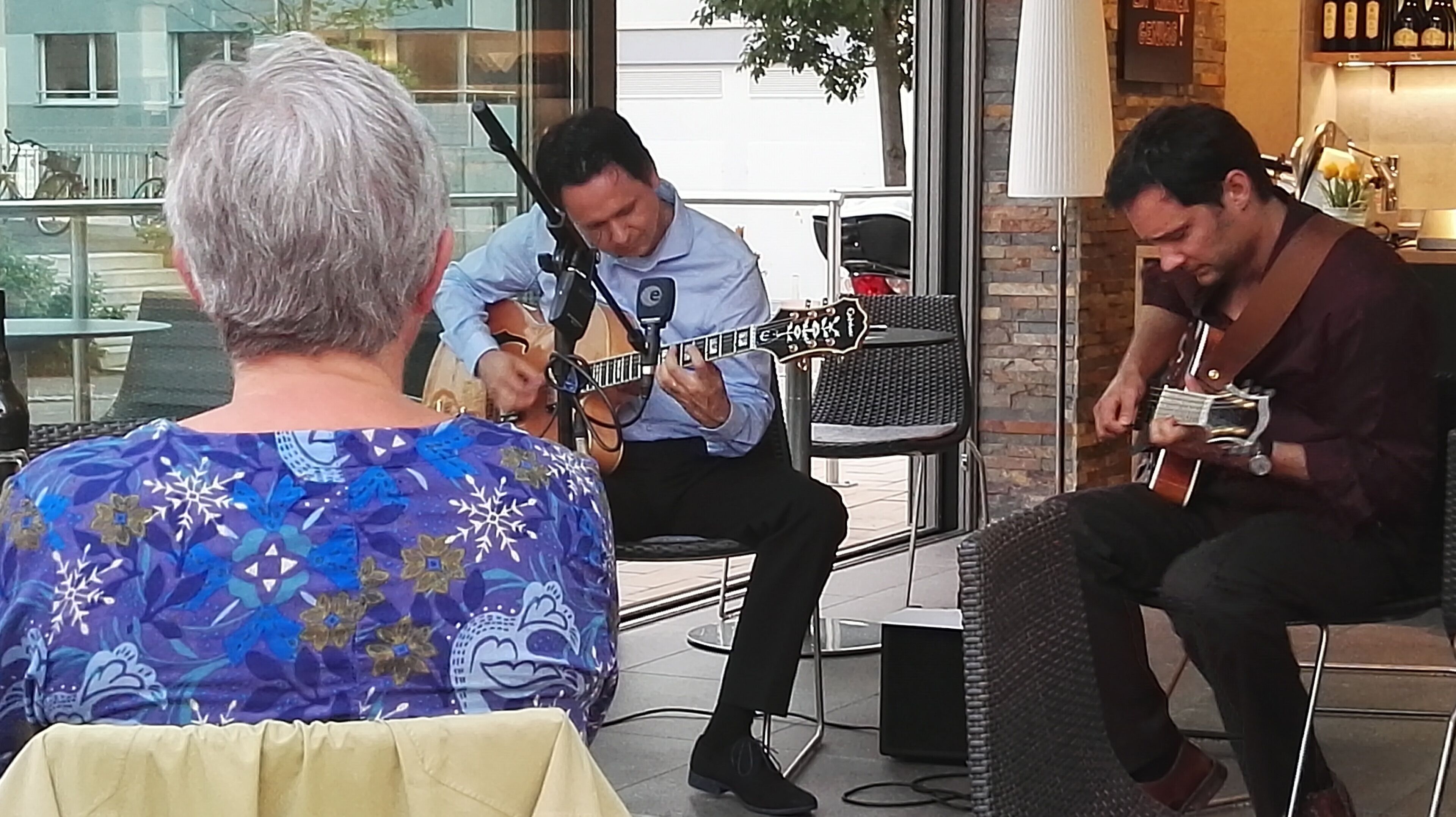 A group of people playing musical instruments outdoors, with a building and a window in the background.