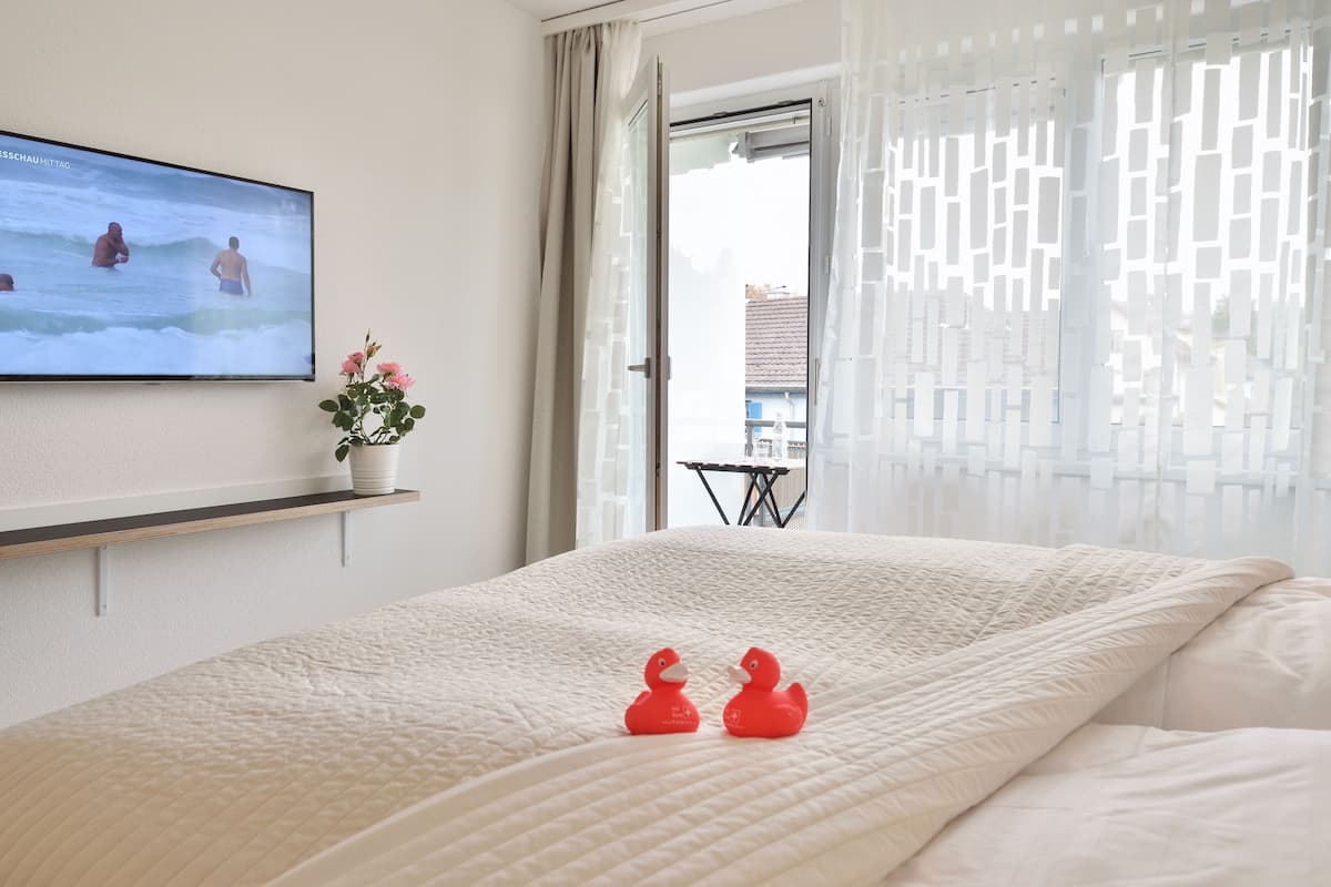 Quarto de hotel com uma cama, uma televisão na parede, uma mesinha com uma planta e vista para o exterior através de uma janela com cortinas leves.