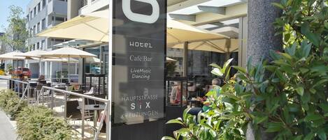 Exterior de un hotel con mesas al aire libre, y un letrero que anuncia servicios como café, bar, música en vivo y baile.