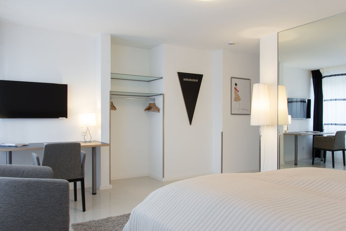 Quarto de hotel moderno com uma cama grande, uma escrivaninha, uma cadeira, uma televisão e um espelho.