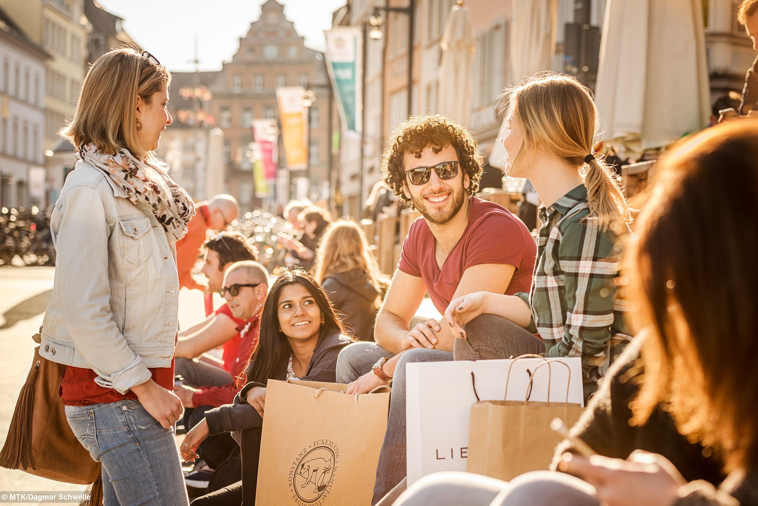 Un grupo de personas sentadas al aire libre, algunas con bolsas de compras.