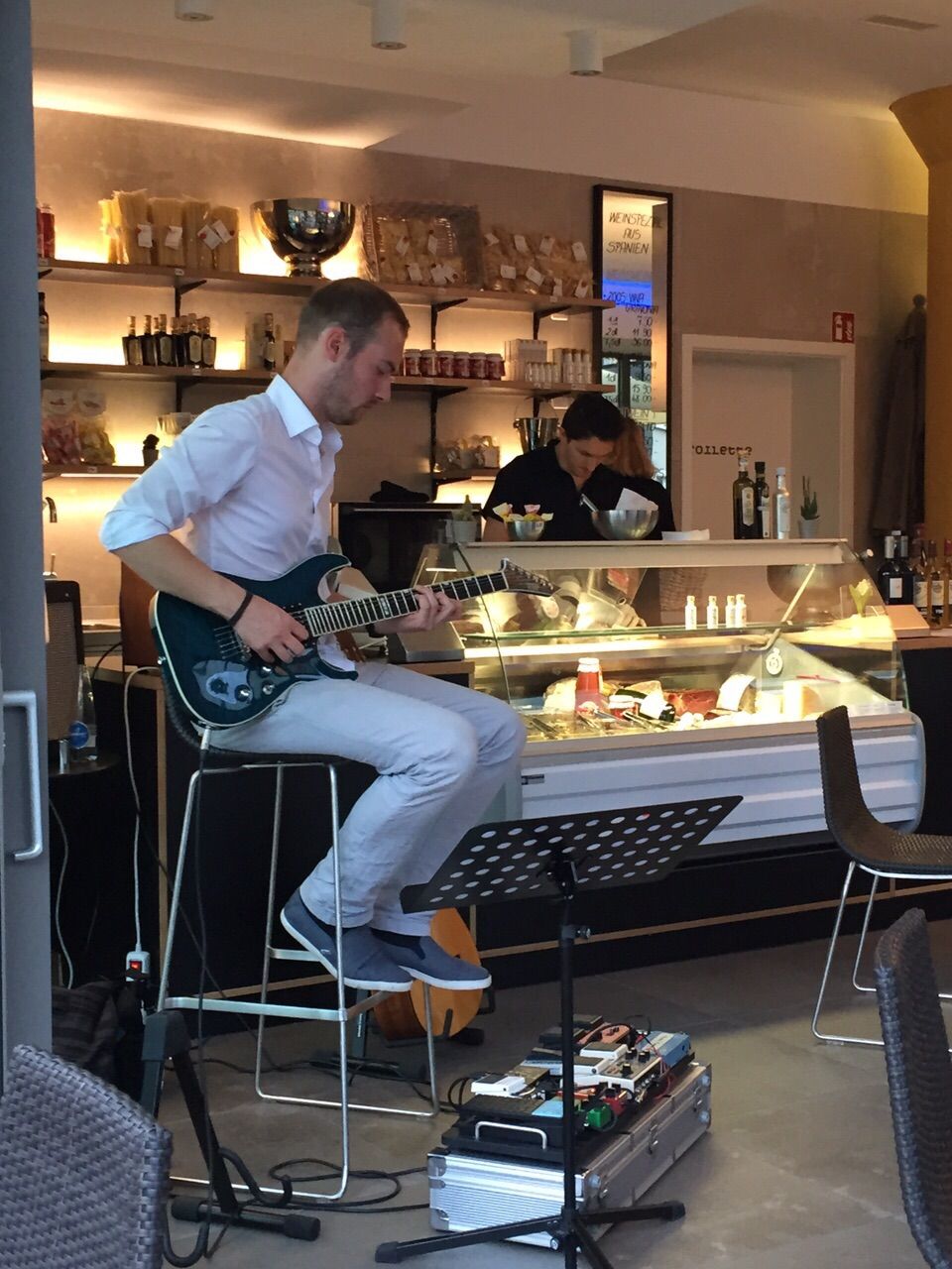 A person playing an electric guitar at a bar area with a counter and shelves stocked with bottles.