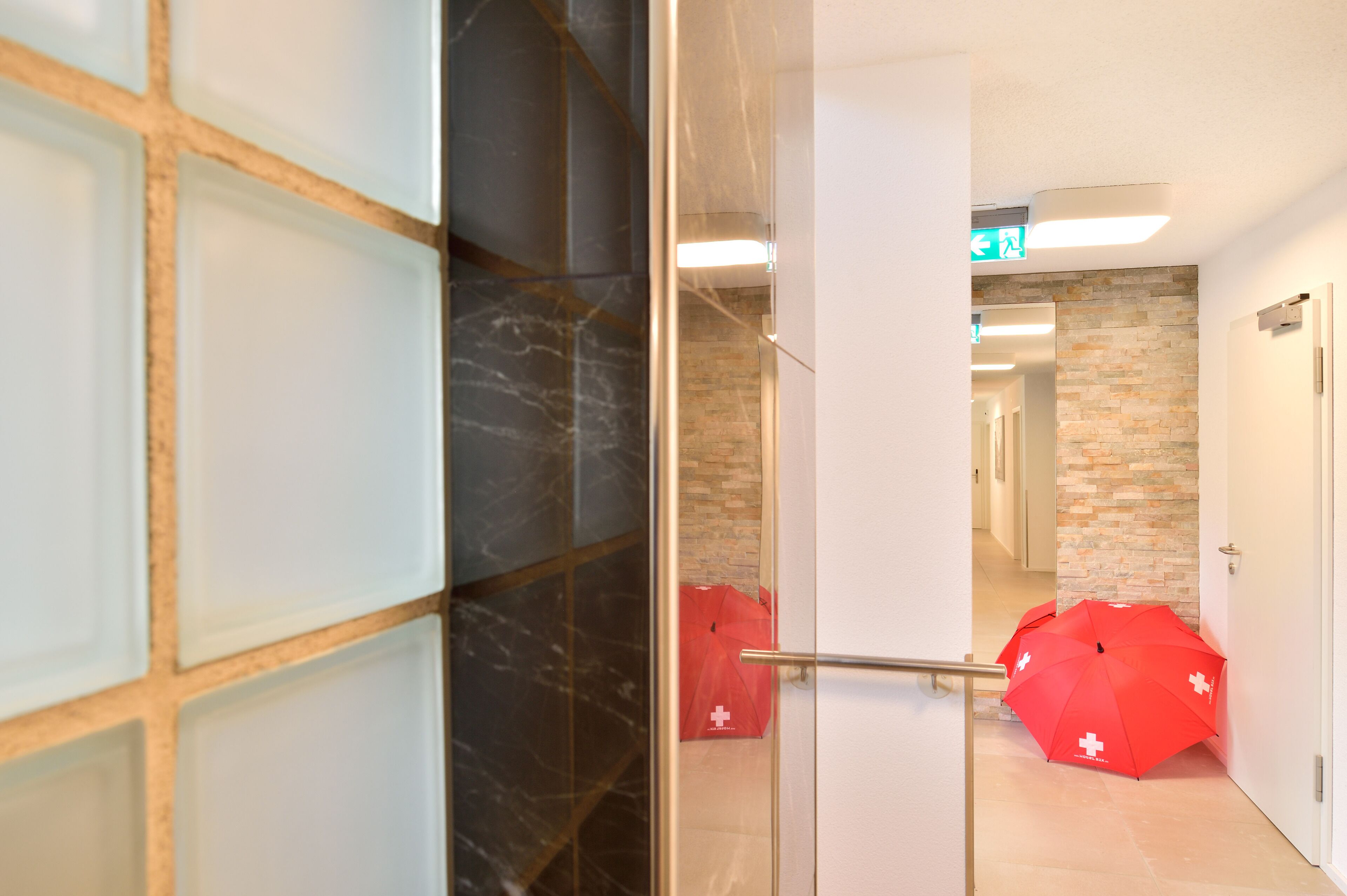 A hallway with a glass door, a marble wall, and two red umbrellas with white crosses.