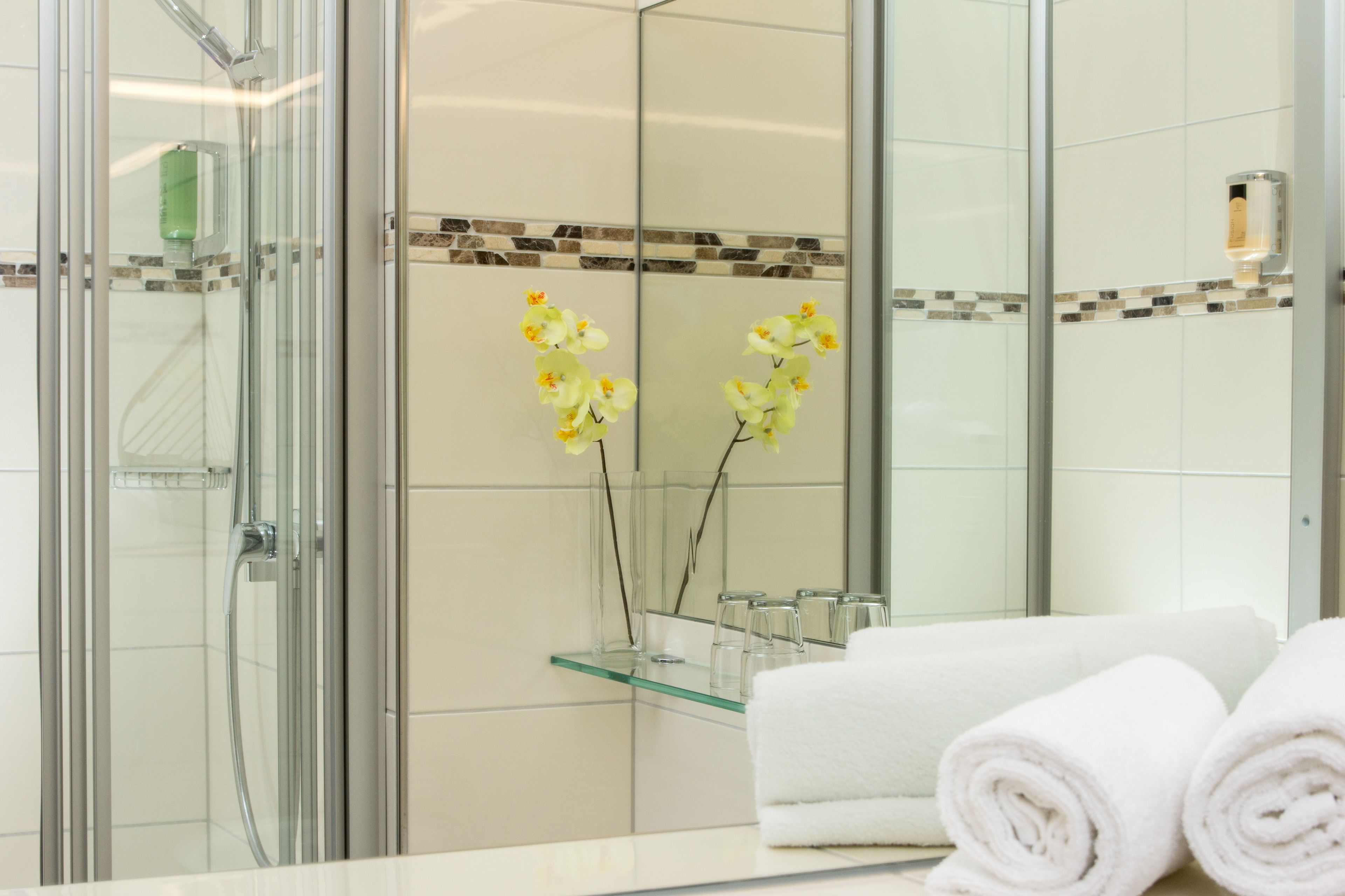 A bathroom with a glass shower enclosure, a glass shelf with toiletries, and a mirror reflecting yellow orchids.