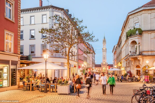 A cobblestone street with outdoor seating, historic buildings, and a prominent tower in the background.