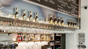 Una zona de bar con un menú en una pizarra, estantes llenos de botellas, vasos y una máquina de café.