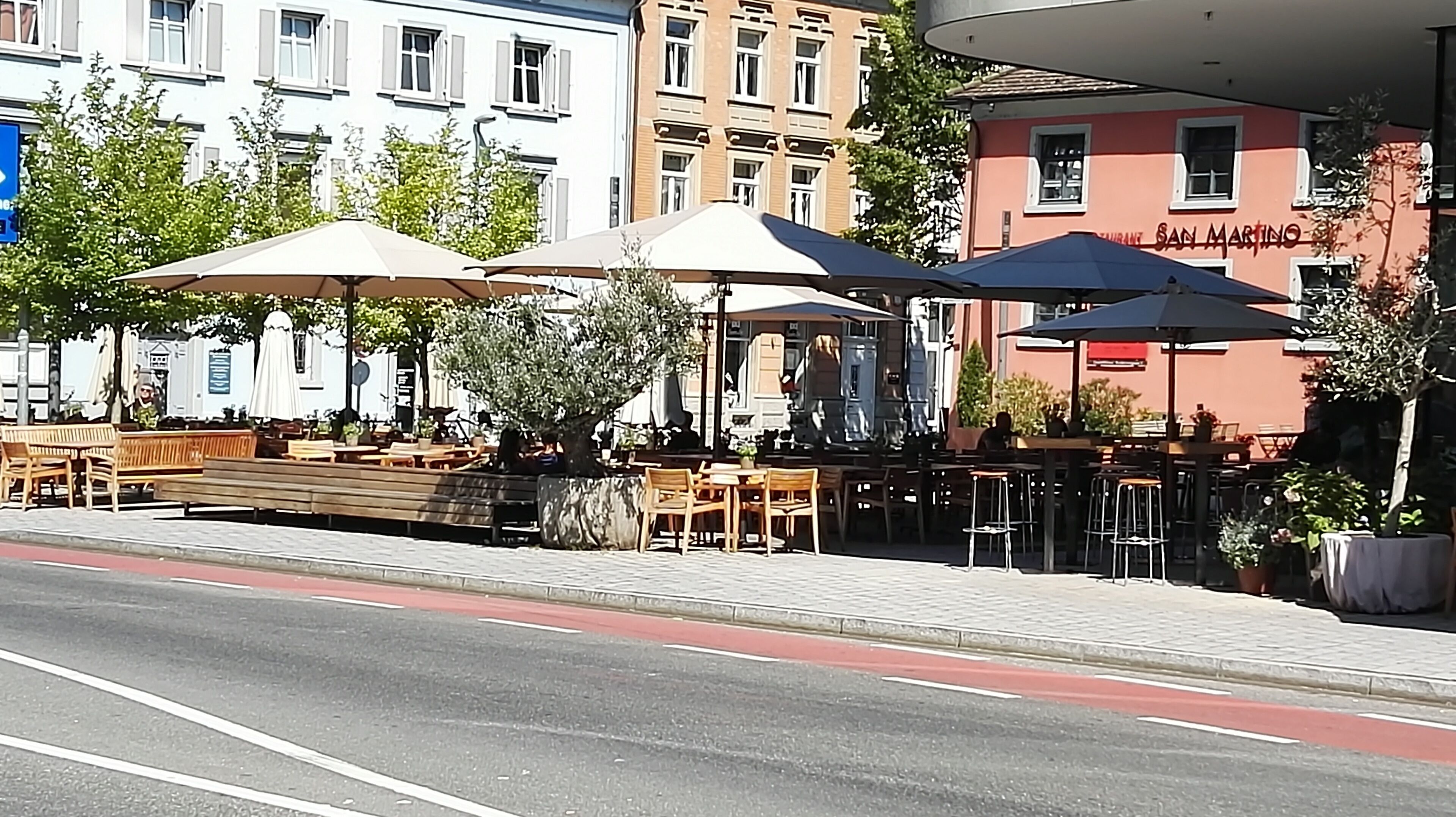 An outdoor café with wooden tables and chairs, umbrellas, and potted plants.