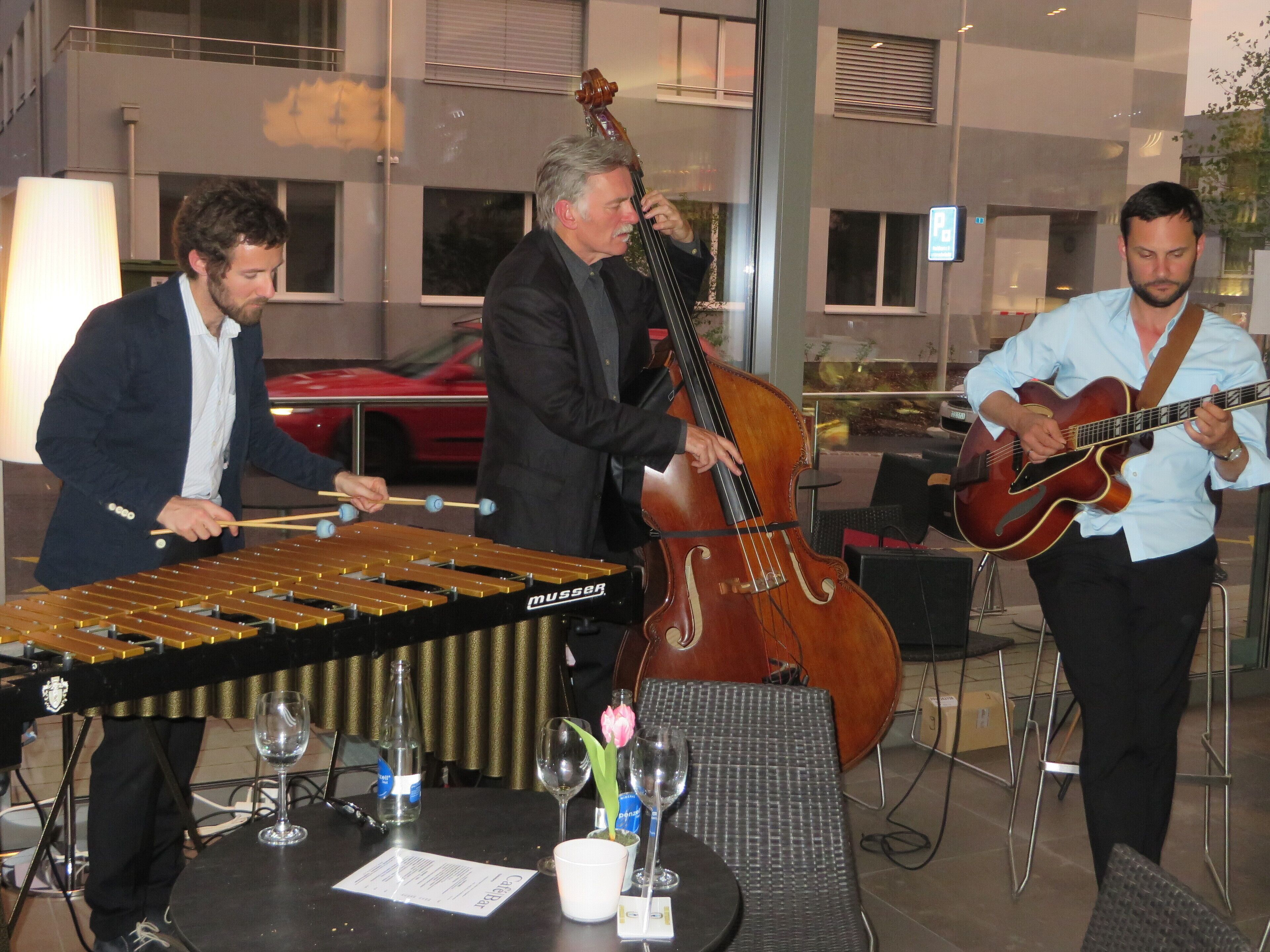 A group of musicians performing in an indoor setting with tables, chairs, and a glass partition.