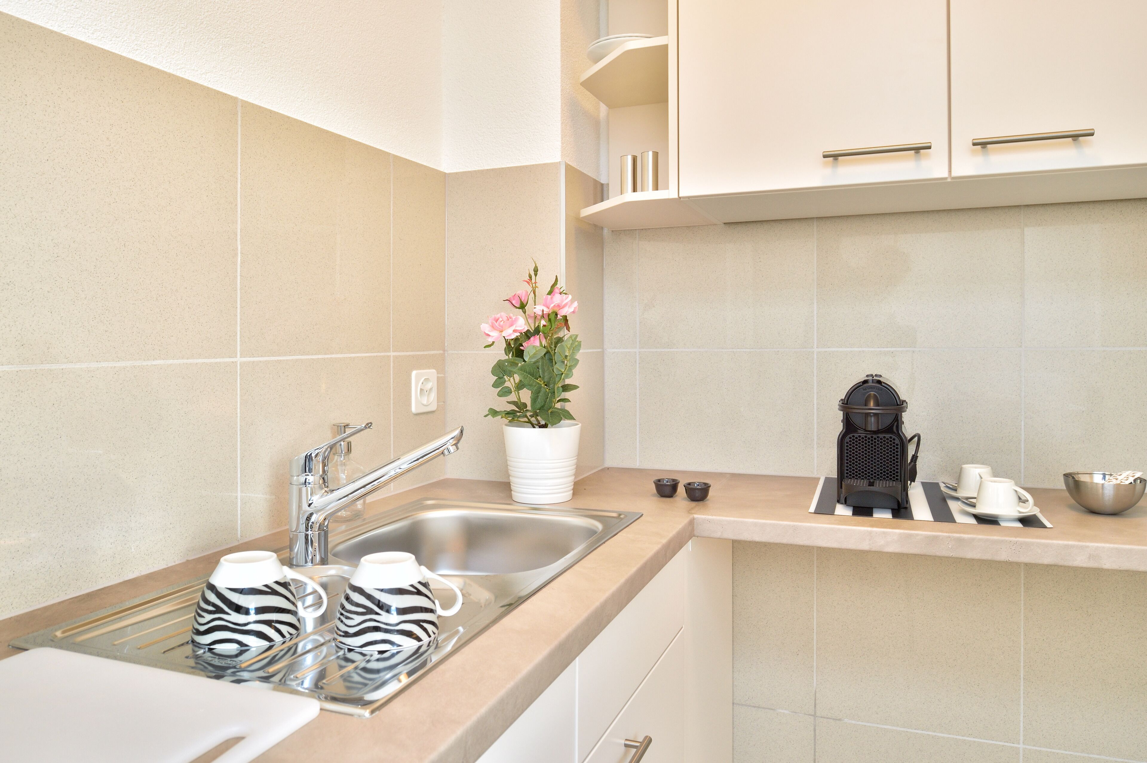 A modern kitchen with a stainless steel sink, two zebra-patterned mugs, a potted plant with pink flowers, a coffee maker, and a bowl on the countertop.