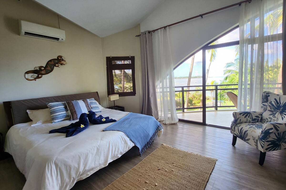 A bedroom with a large bed, a wooden dresser, a chair, and a balcony with a view of greenery.
