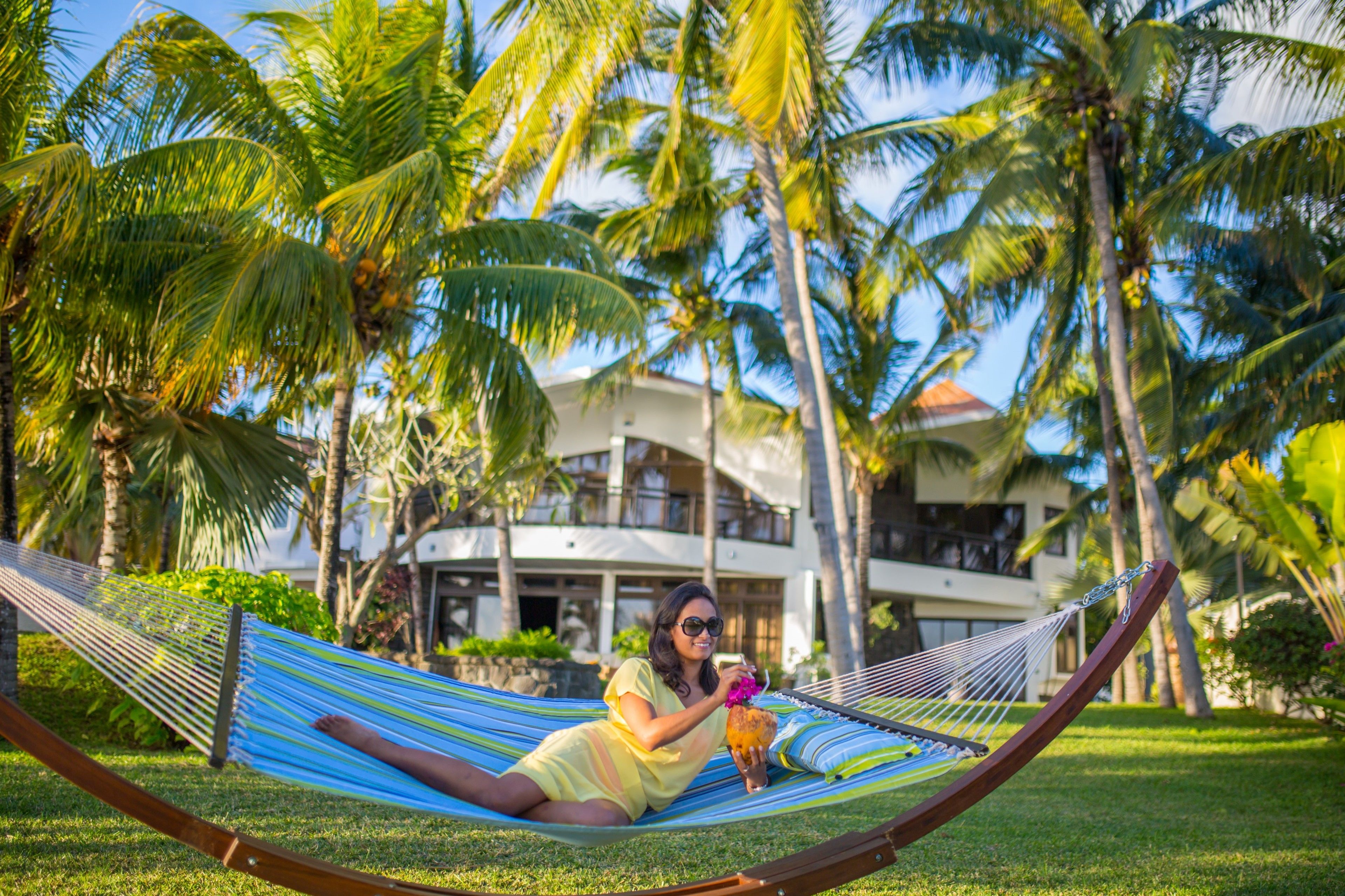 A person relaxing on a hammock in a lush, tropical setting with palm trees and a large building in the background.