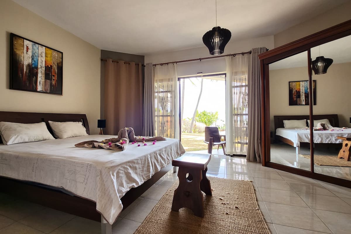 A spacious bedroom with a large bed, a small table, and a chair. There is a sliding glass door leading to an outdoor area.