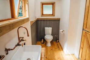 Een kleine badkamer met een houten vloer, een witte wastafel met koperen details, een toilet en een klein raam boven het toilet.