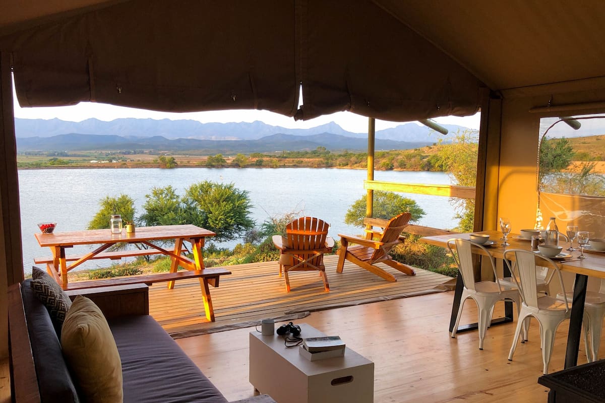 Een houten terras met een tafel en stoelen met uitzicht op een meer, met bergen op de achtergrond.