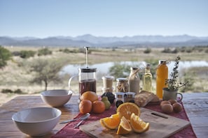 Un montaje para picnic con una mesa de madera, cuencos, naranjas, una tabla de cortar y vistas a montañas y agua al fondo.