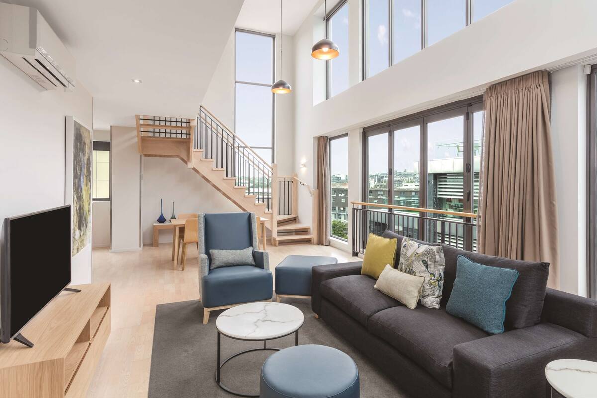 Ein großzügiger Wohnbereich mit Sofa, Hocker und TV-Möbel, einer Treppe und großen Fenstern mit Blick auf die Stadt.