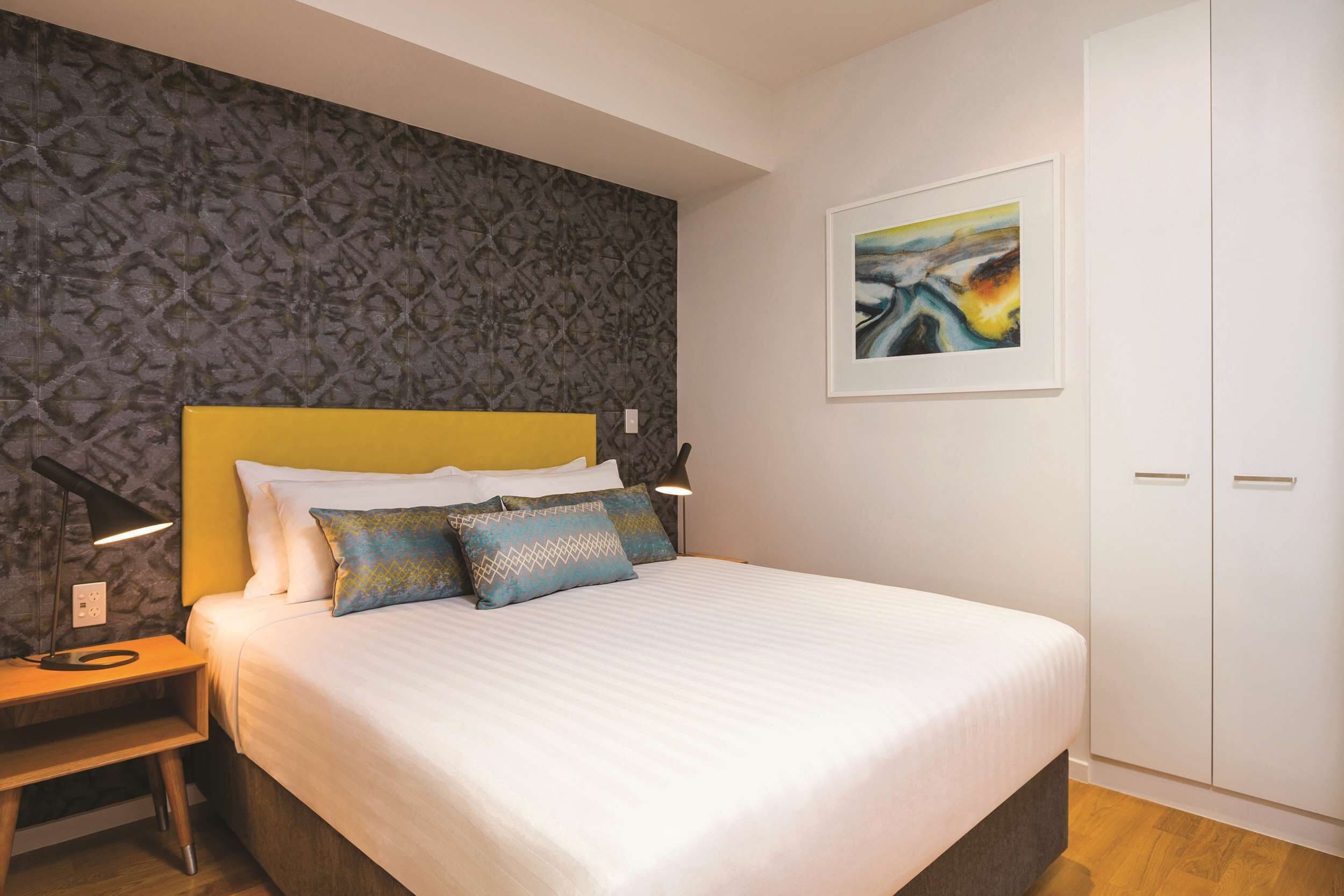 A neatly made bed with white linens and patterned pillows, a bedside lamp, and a wall-mounted artwork.