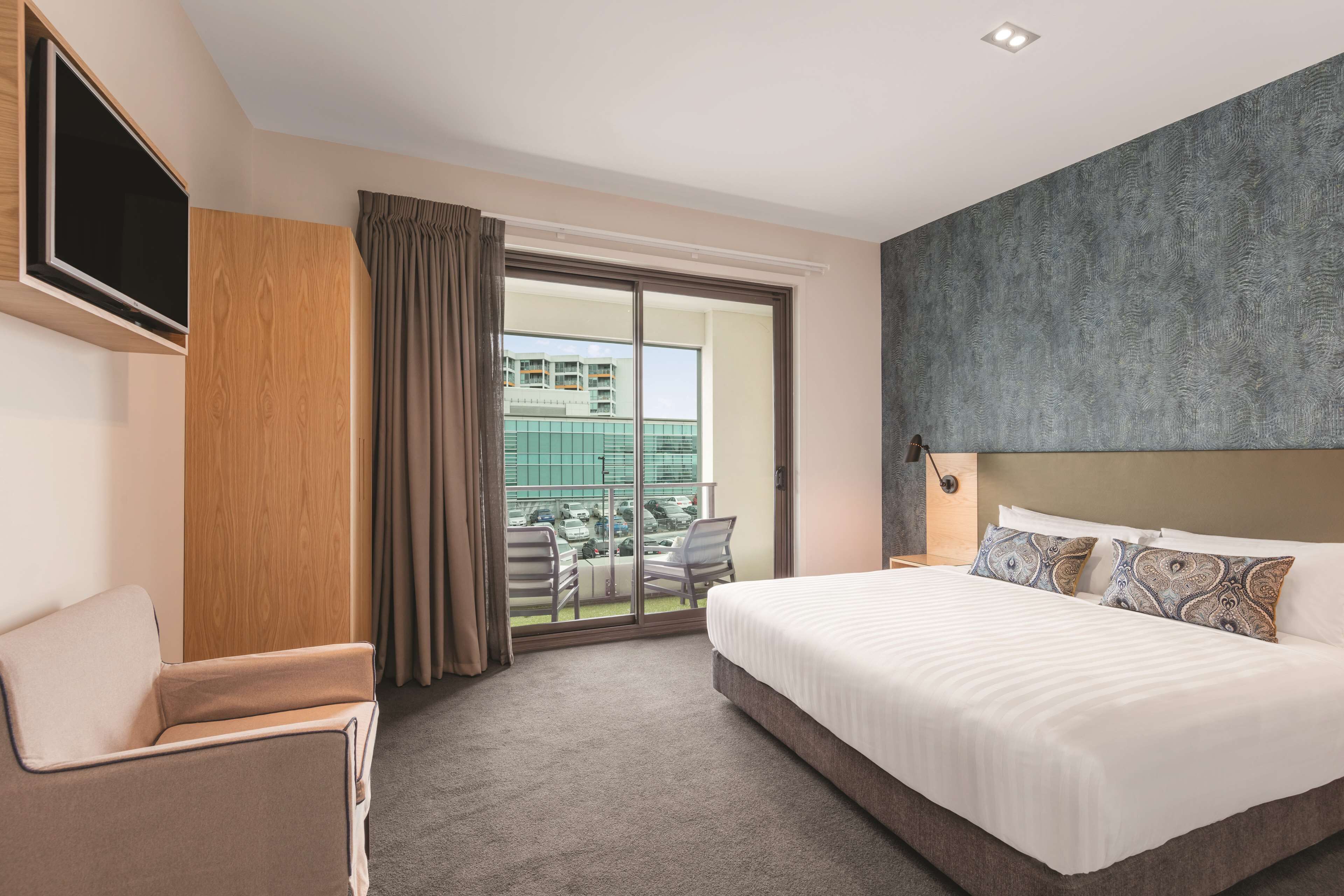 Quarto de hotel com uma cama grande, televisão de tela plana na parede, área de estar com uma poltrona e uma varanda com vista para a cidade.