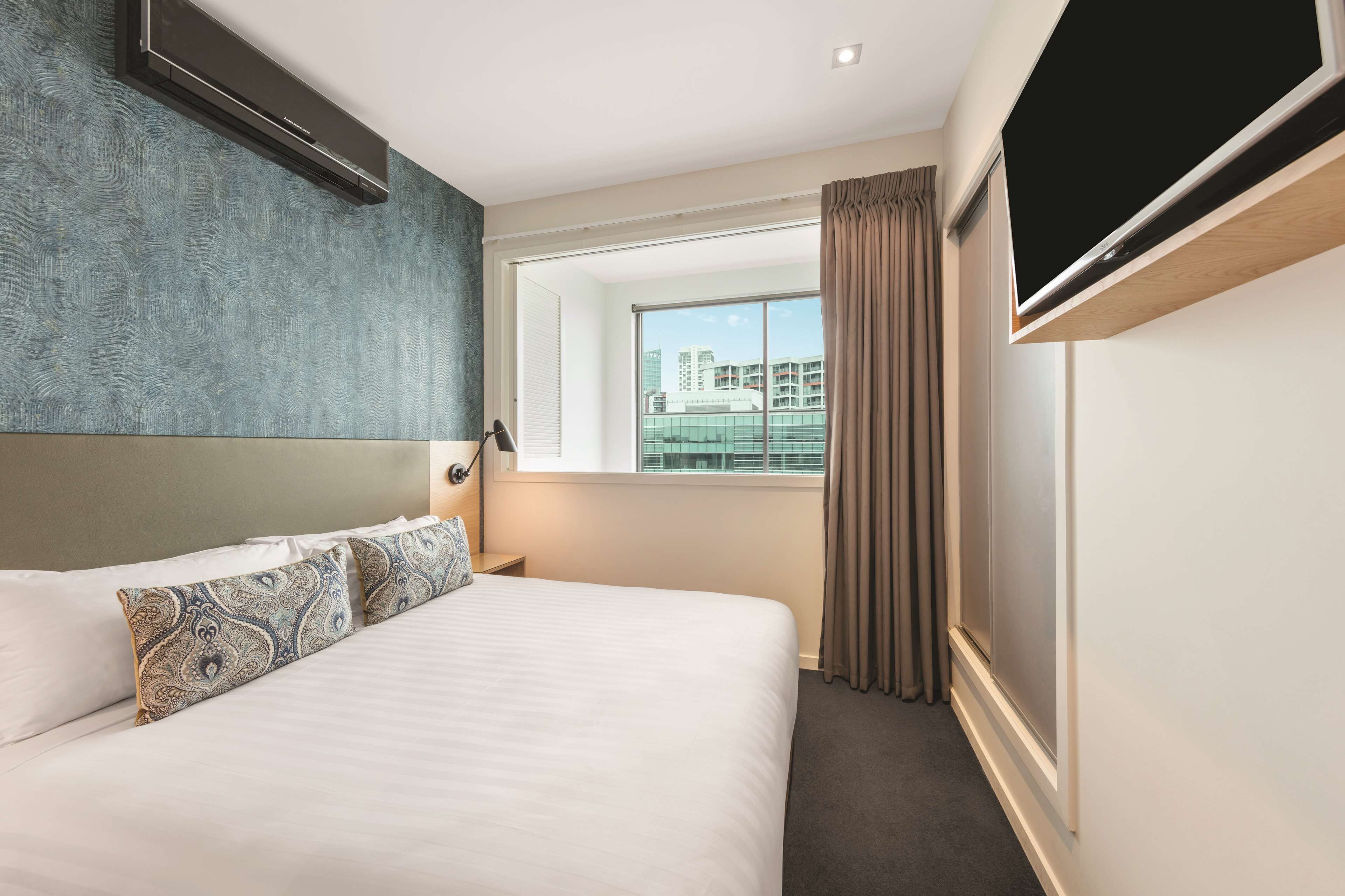 Une chambre d’hôtel avec un grand lit, un téléviseur fixé au mur, une fenêtre avec des rideaux et une vue sur les bâtiments environnants.