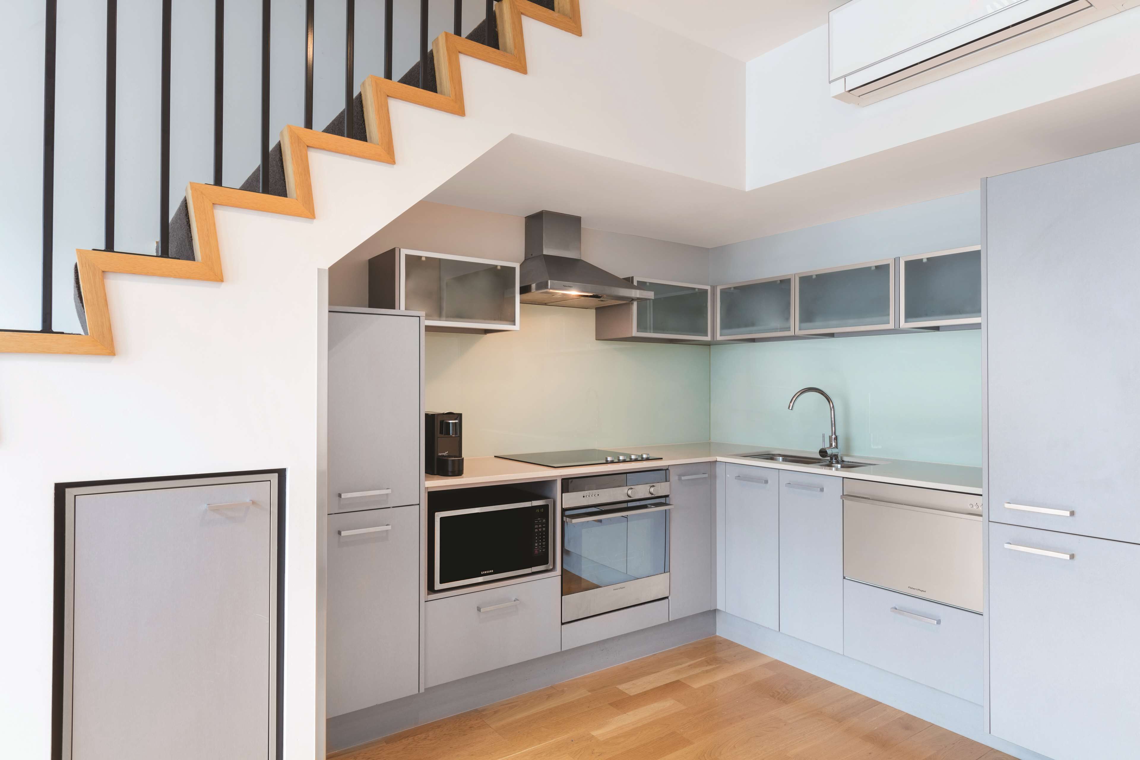 A modern kitchen with a built-in oven, microwave, and dishwasher, a wooden staircase, and a light-colored floor.
