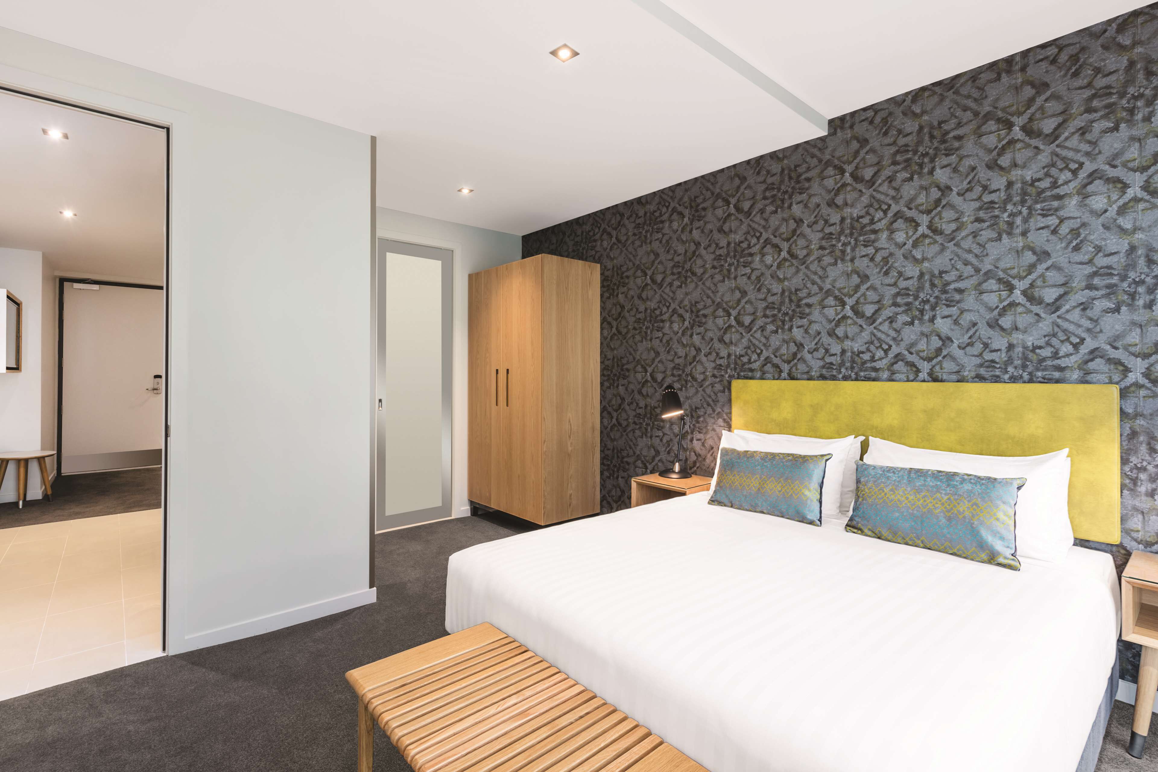 A modern hotel room with a large bed, a wooden bench, a wardrobe, and a patterned wall.