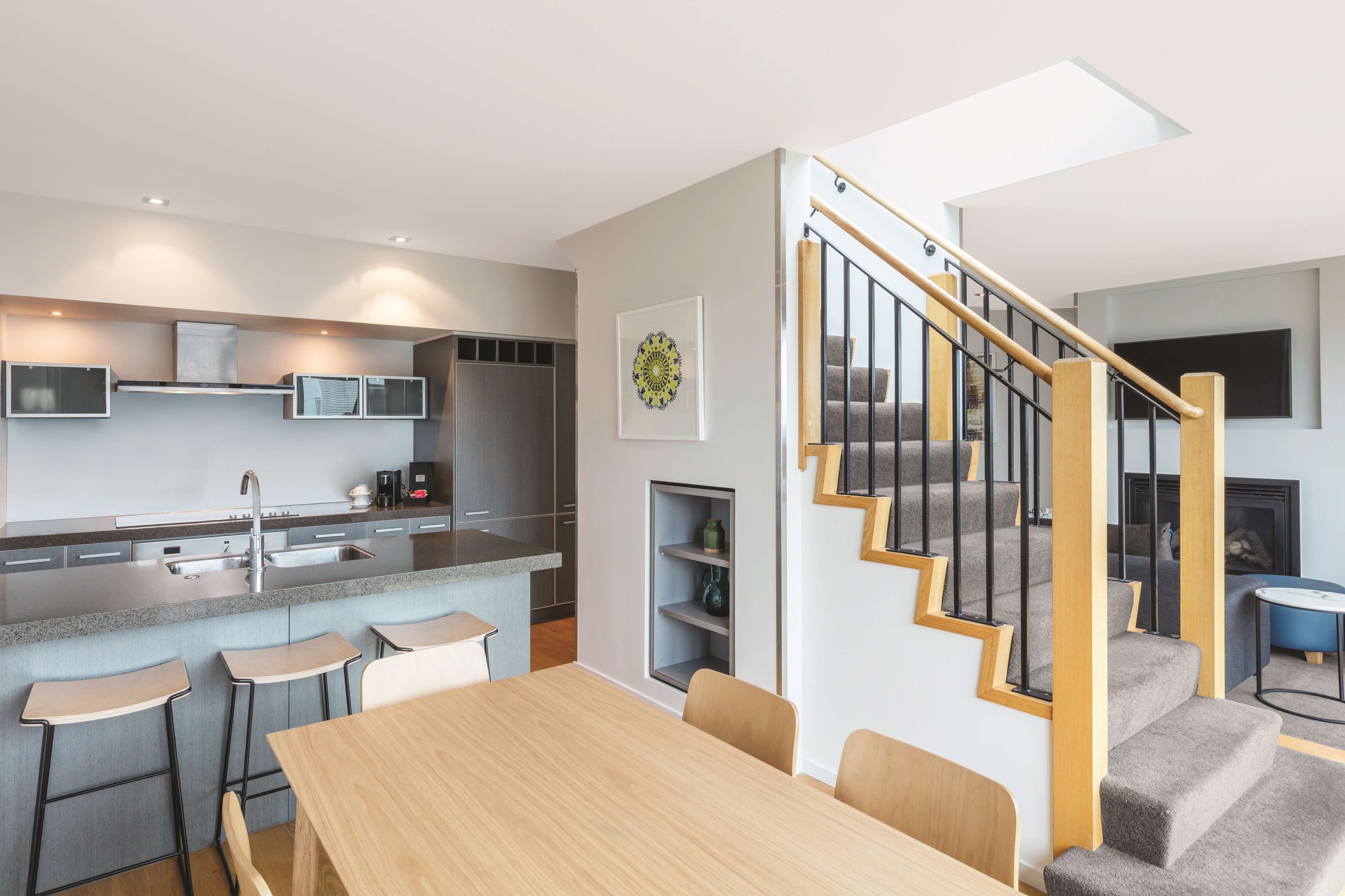 Une cuisine moderne dotée d’un îlot central, de tabourets de bar, d’une table à manger et d’un escalier avec une rampe en verre.