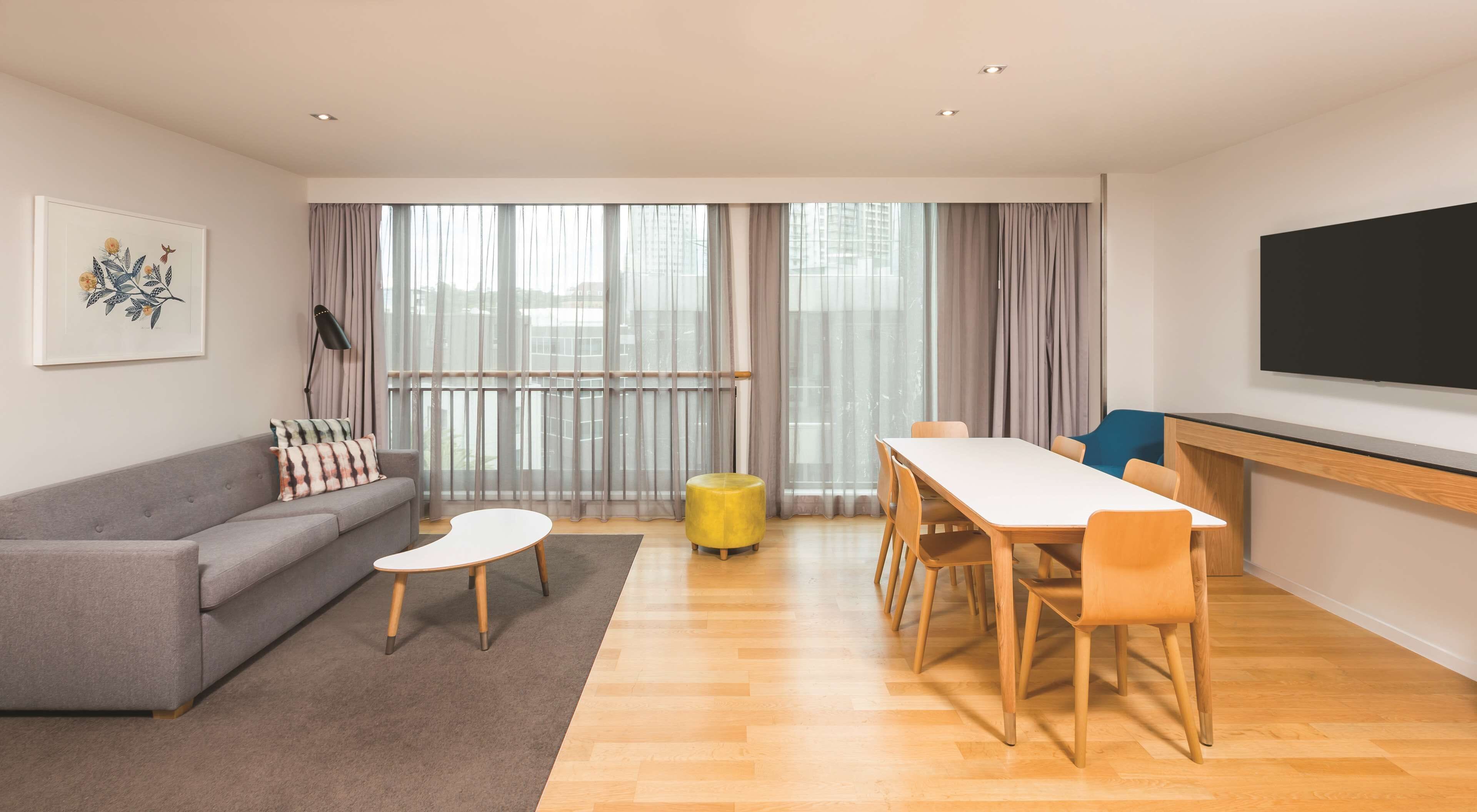 Una sala de estar moderna con un sofá gris, una mesita de centro blanca, un reposapiés amarillo y una mesa de comedor de madera con sillas. Hay un ventanal con cortinas translúcidas, una pizarra en la pared y un cuadro enmarcado sobre el sofá.