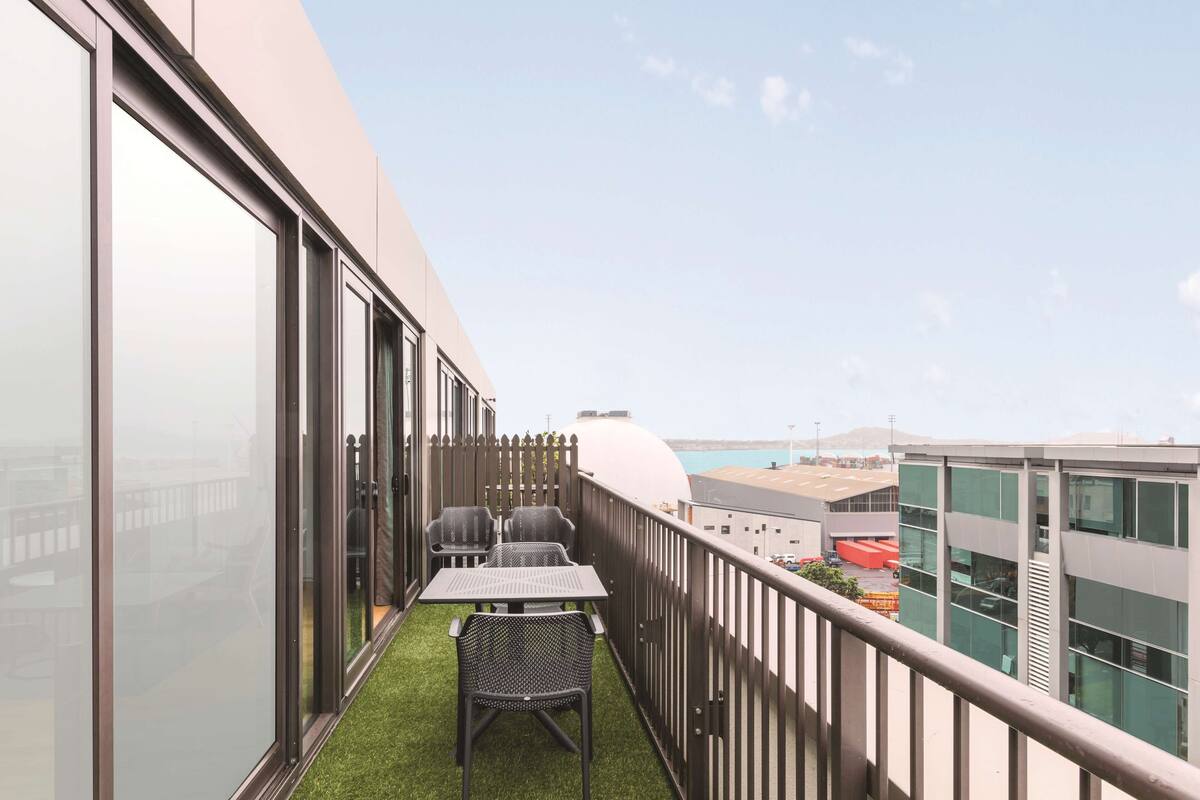 Ein Balkon mit Tisch und Stühlen, einer kleinen Fläche aus Kunstrasen und Blick auf Gebäude sowie ein Gewässer im Hintergrund.