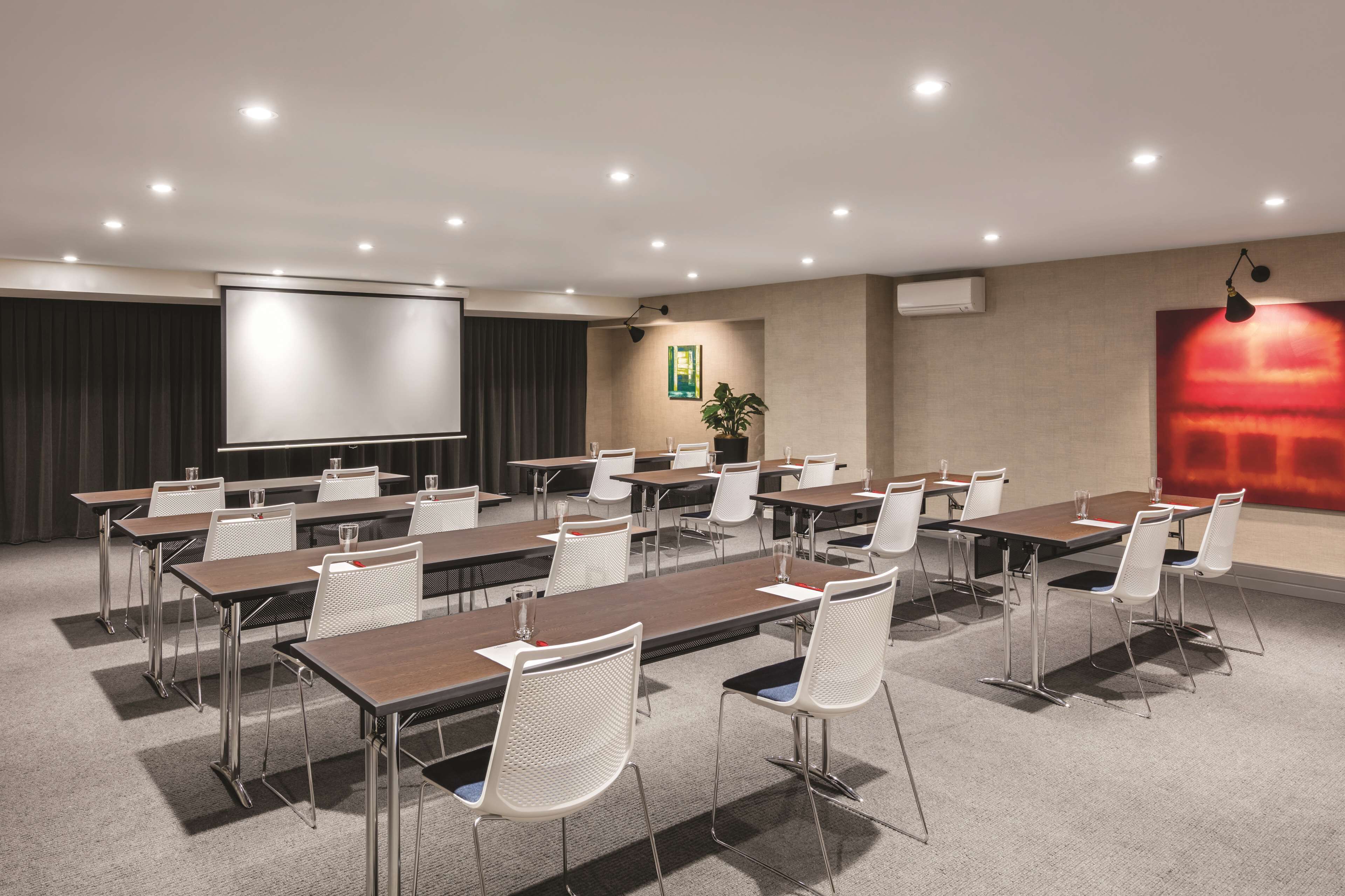 Un salón de conferencias con varias mesas y sillas, una pantalla de proyector y un cuadro abstracto grande de color rojo en la pared.