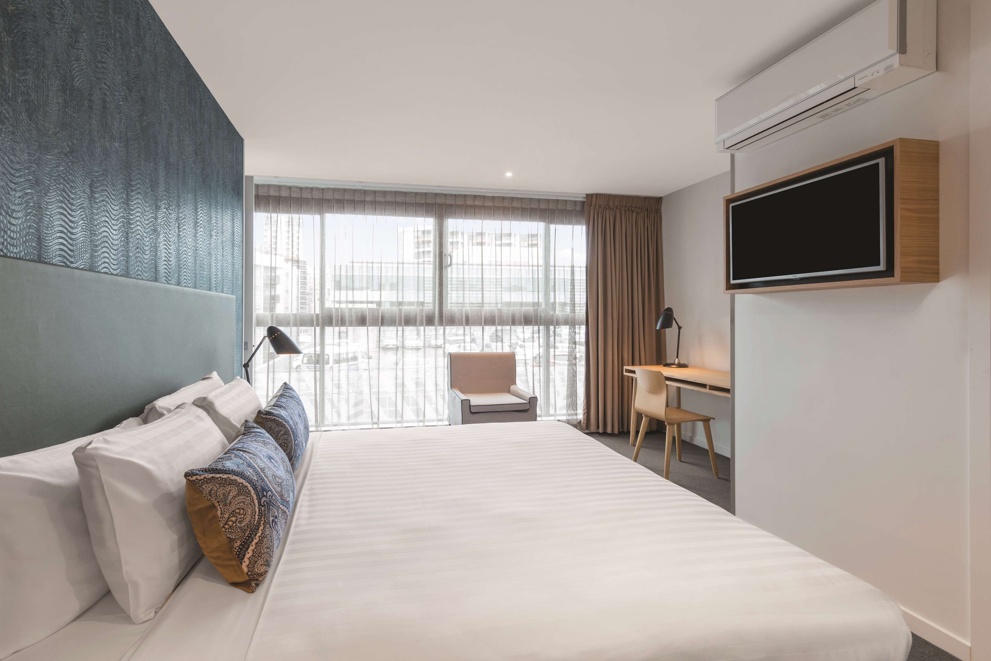 A modern hotel room with a large bed, a desk, a chair, and a TV mounted on the wall.
