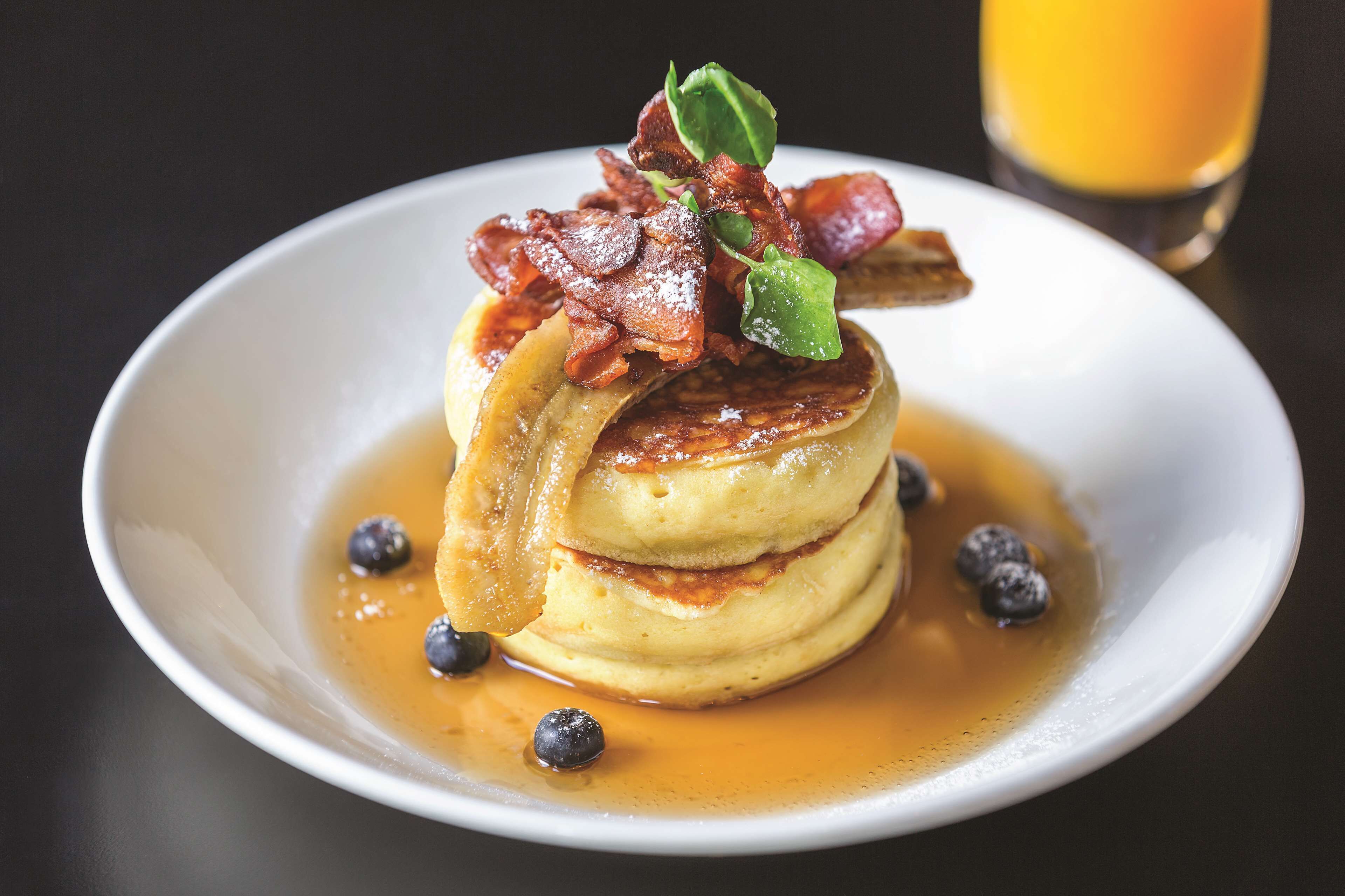 A stack of pancakes with syrup, bacon, and blueberries on a white plate.