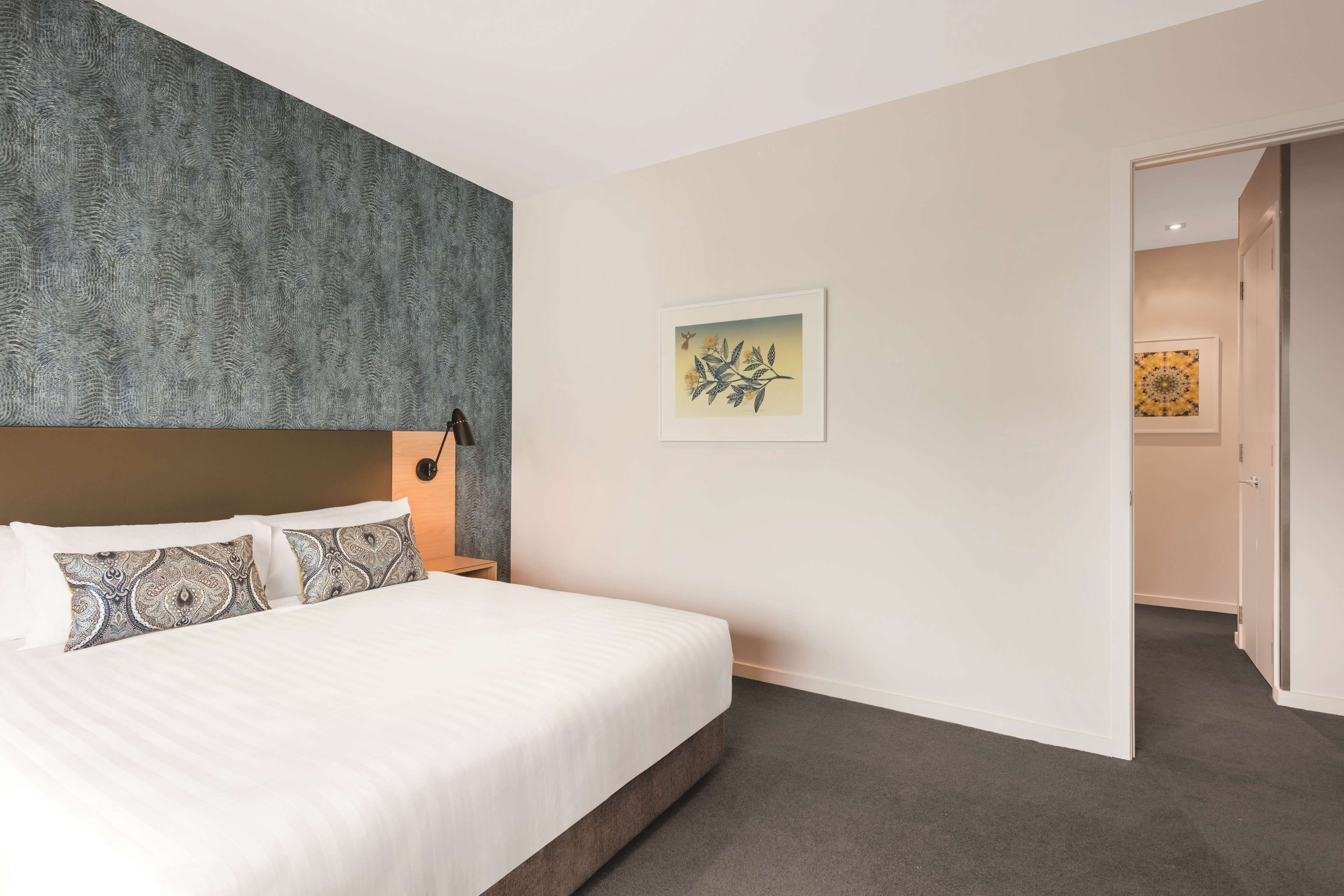 A hotel room with a large bed, a headboard with a textured design, a wall-mounted lamp, and a framed artwork.