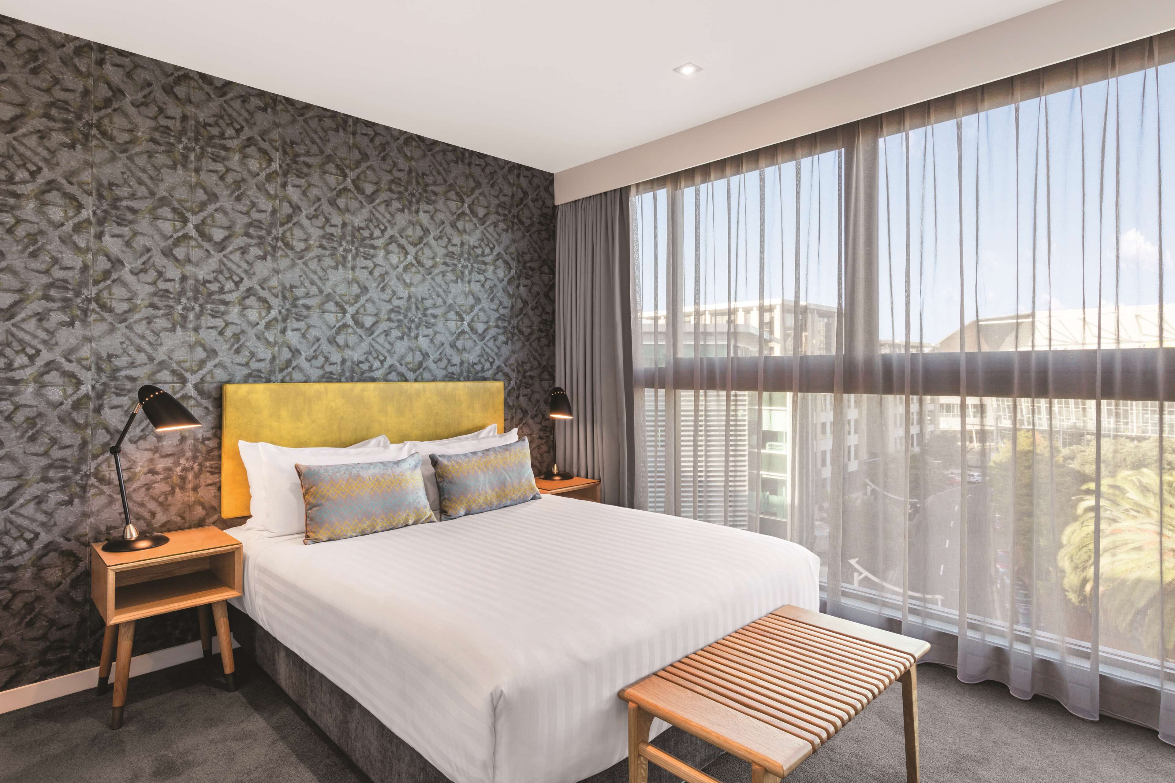 A modern hotel room with a large bed, two bedside tables with lamps, a bench, and a view of the city through sheer curtains.