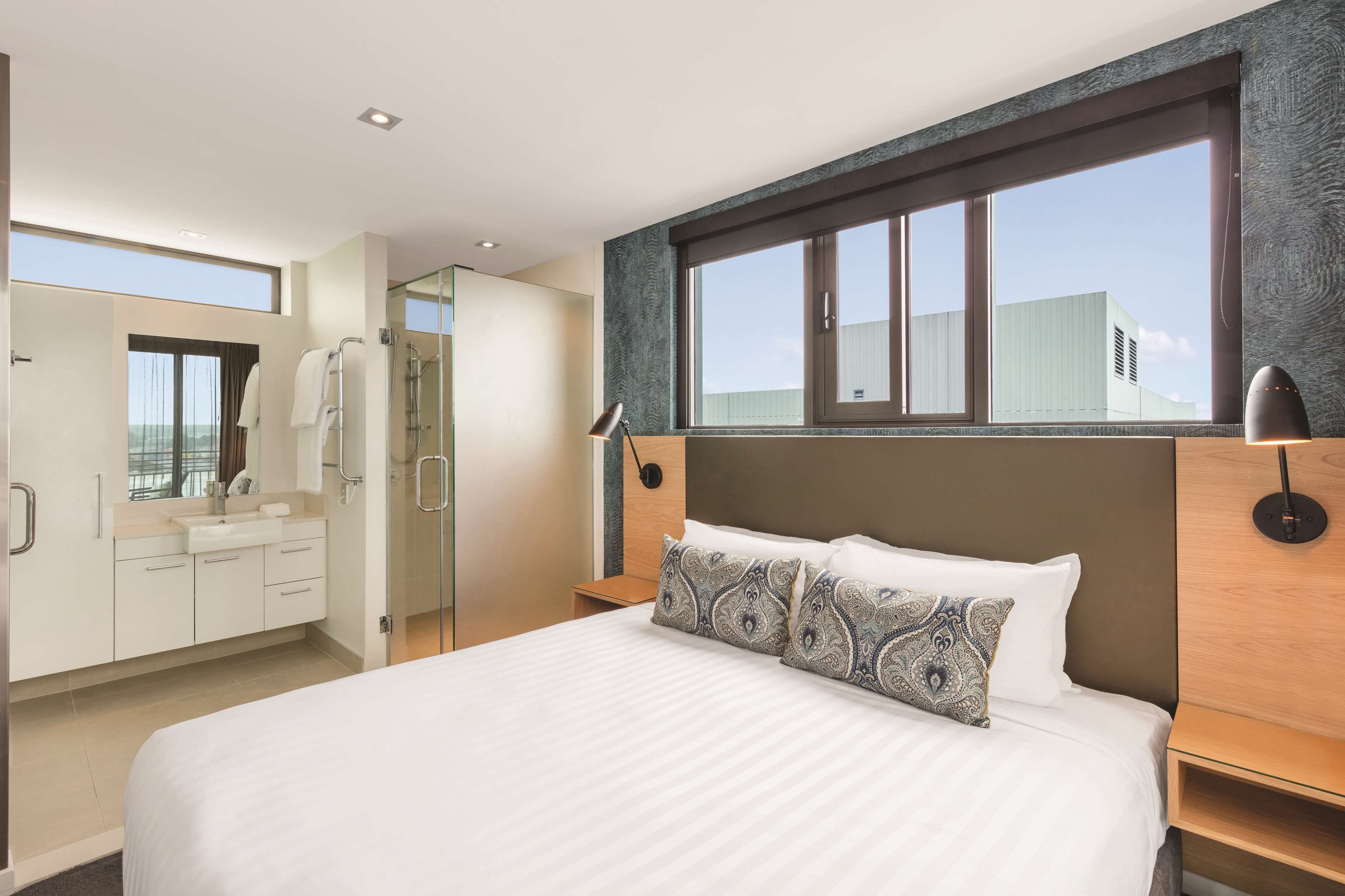 A modern hotel room with a large bed, a walk-in closet, a shower area, and a large window offering a city view.