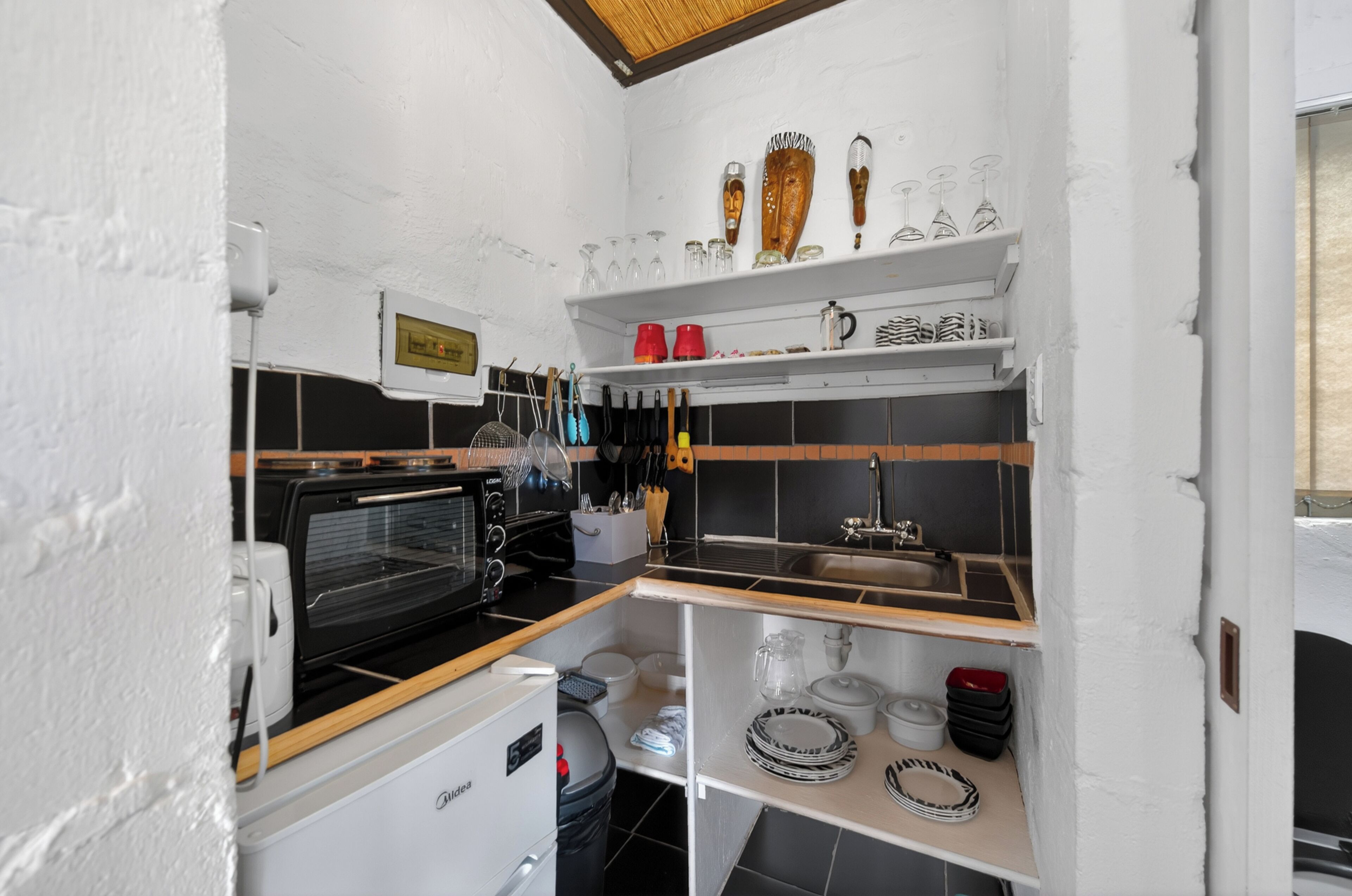 Cocina compacta con microondas, horno y lavamanos. Se observan estantes con cristalería y utensilios.