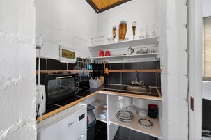 Cocina compacta con microondas, horno y lavamanos. Se observan estantes con cristalería y utensilios.