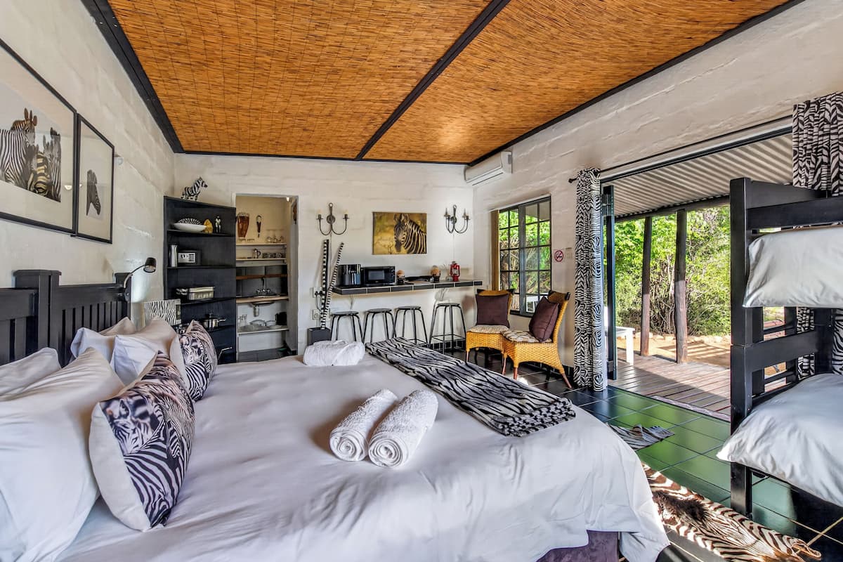 A room with a large bed, a kitchenette, a dining area, and a view of greenery.