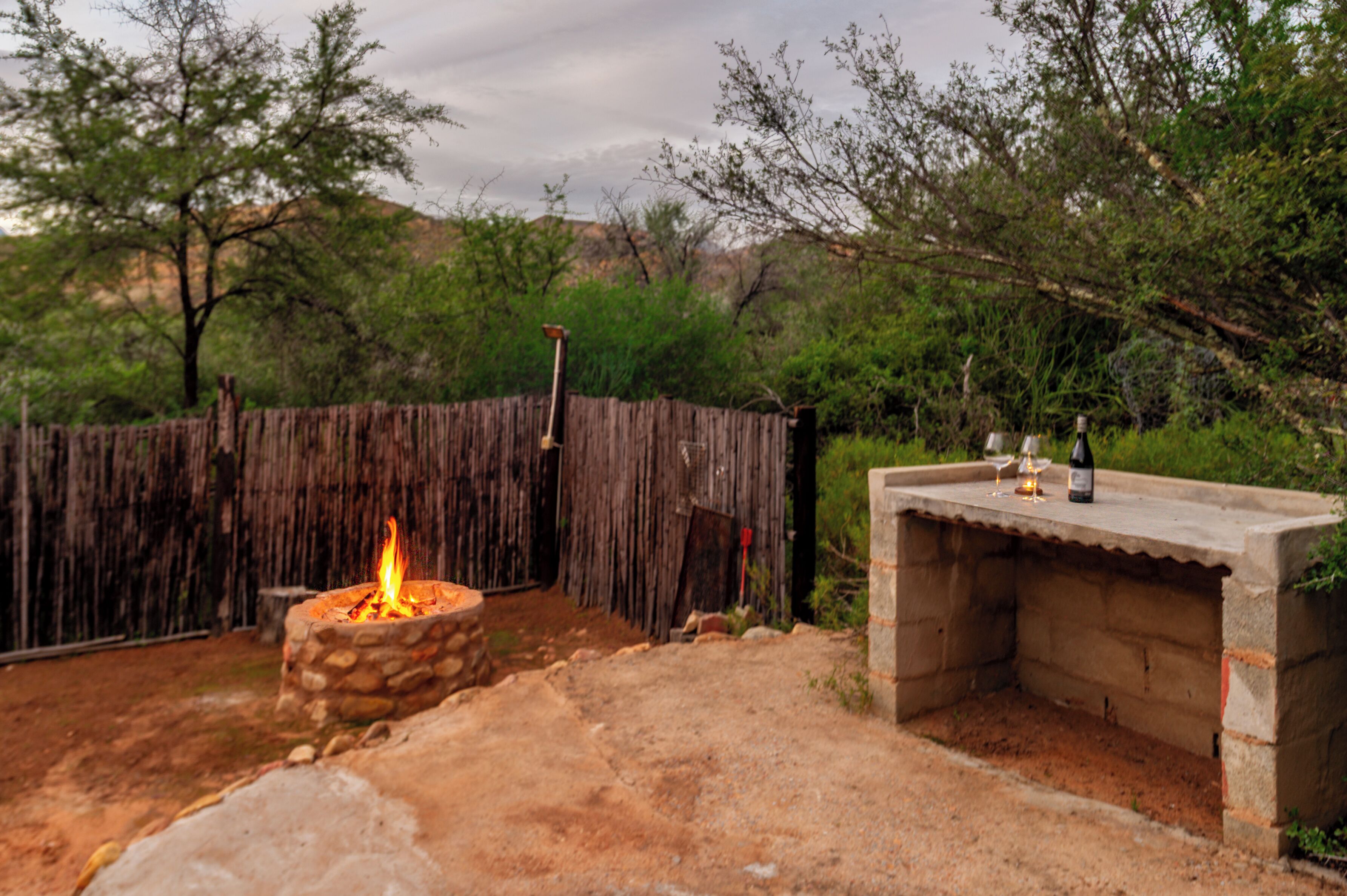 A stone fire pit with a flame, a concrete outdoor seating area, and a wooden fence in a natural setting.