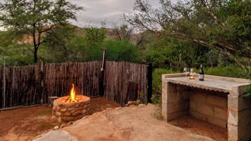 A stone fire pit with a flame, a concrete outdoor seating area, and a wooden fence in a natural setting.