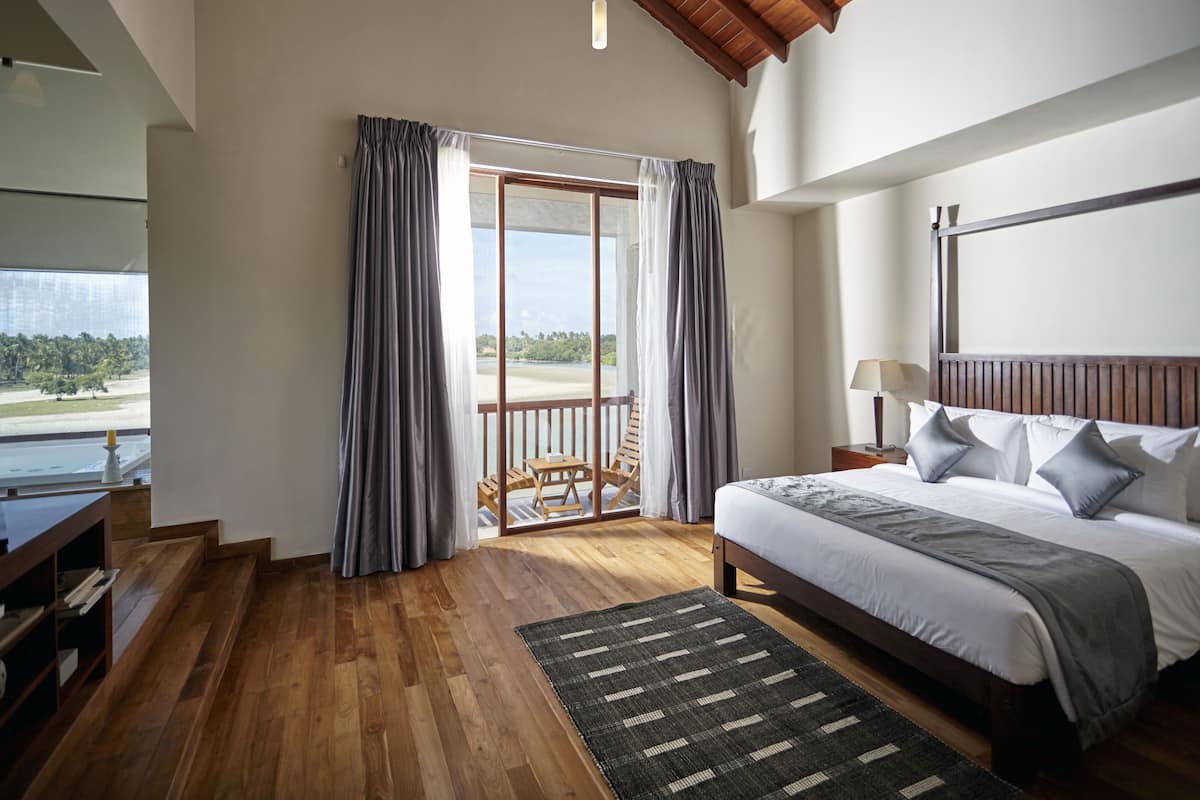 Ein Schlafzimmer mit einem großen Bett, Holzboden, Blick auf einen Strand und einem Balkon mit Vorhängen.