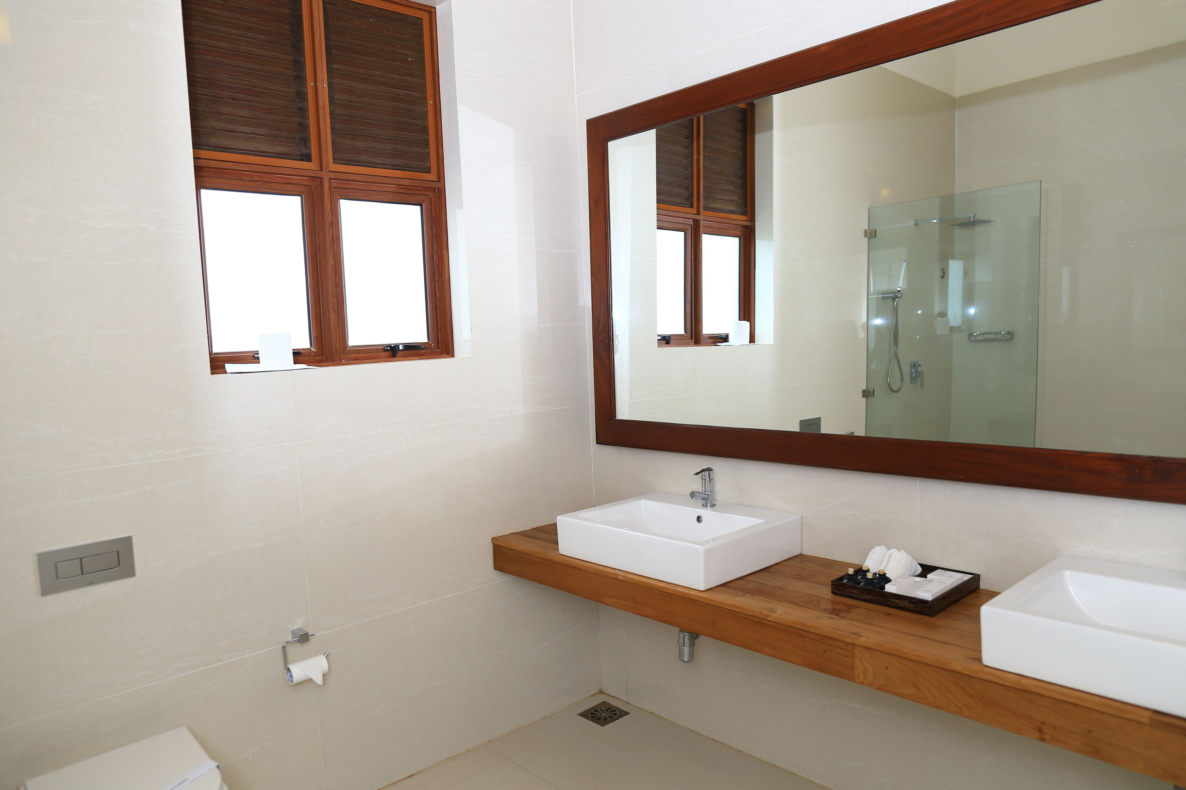 A modern bathroom with a wooden vanity, two sinks, a large mirror, and a window with blinds.