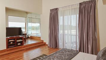 Un dormitorio con una cama grande, televisor, piso de madera y vistas al exterior a través de una ventana con cortinas.