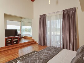 Un dormitorio con una cama grande, televisor, piso de madera y vistas al exterior a través de una ventana con cortinas.