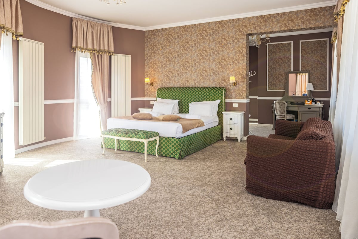 A hotel room with a green bed, white bedside tables, a round white table, and a brown armchair.